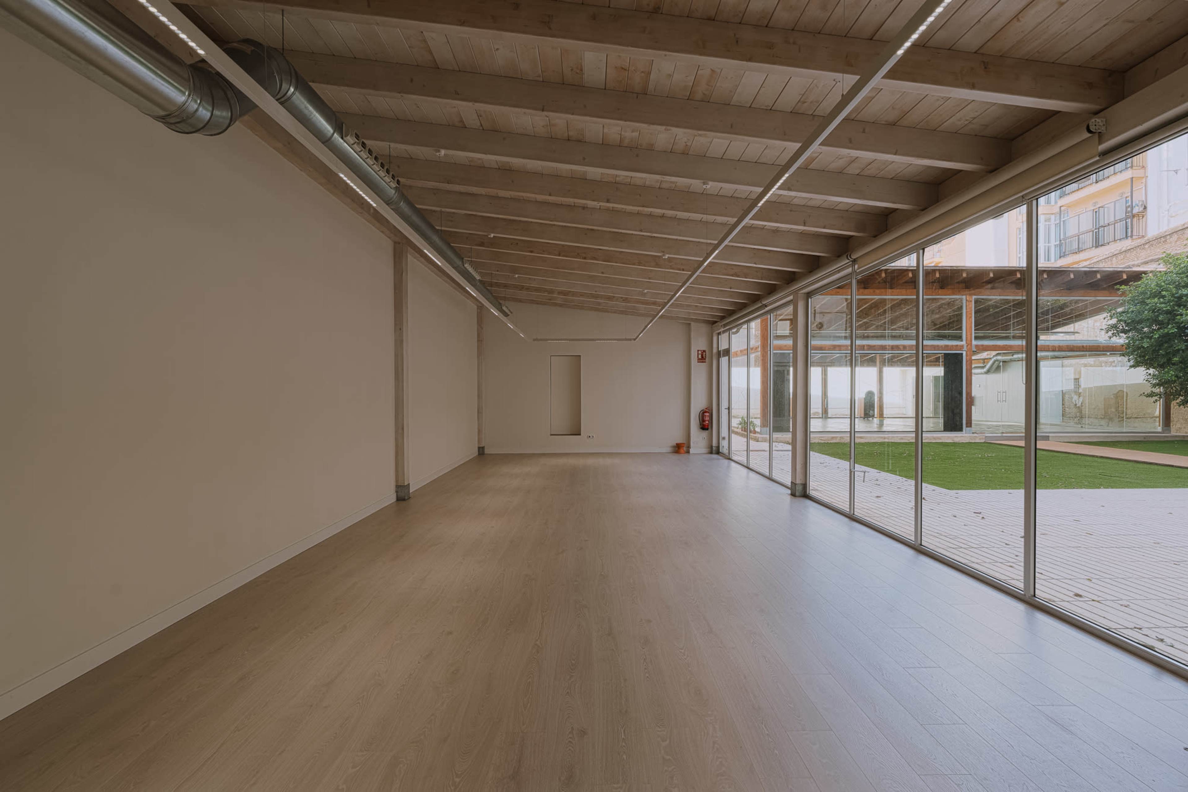 The image shows a spacious, empty room with wooden beams on the ceiling, large glass windows, and a view of an outdoor area.