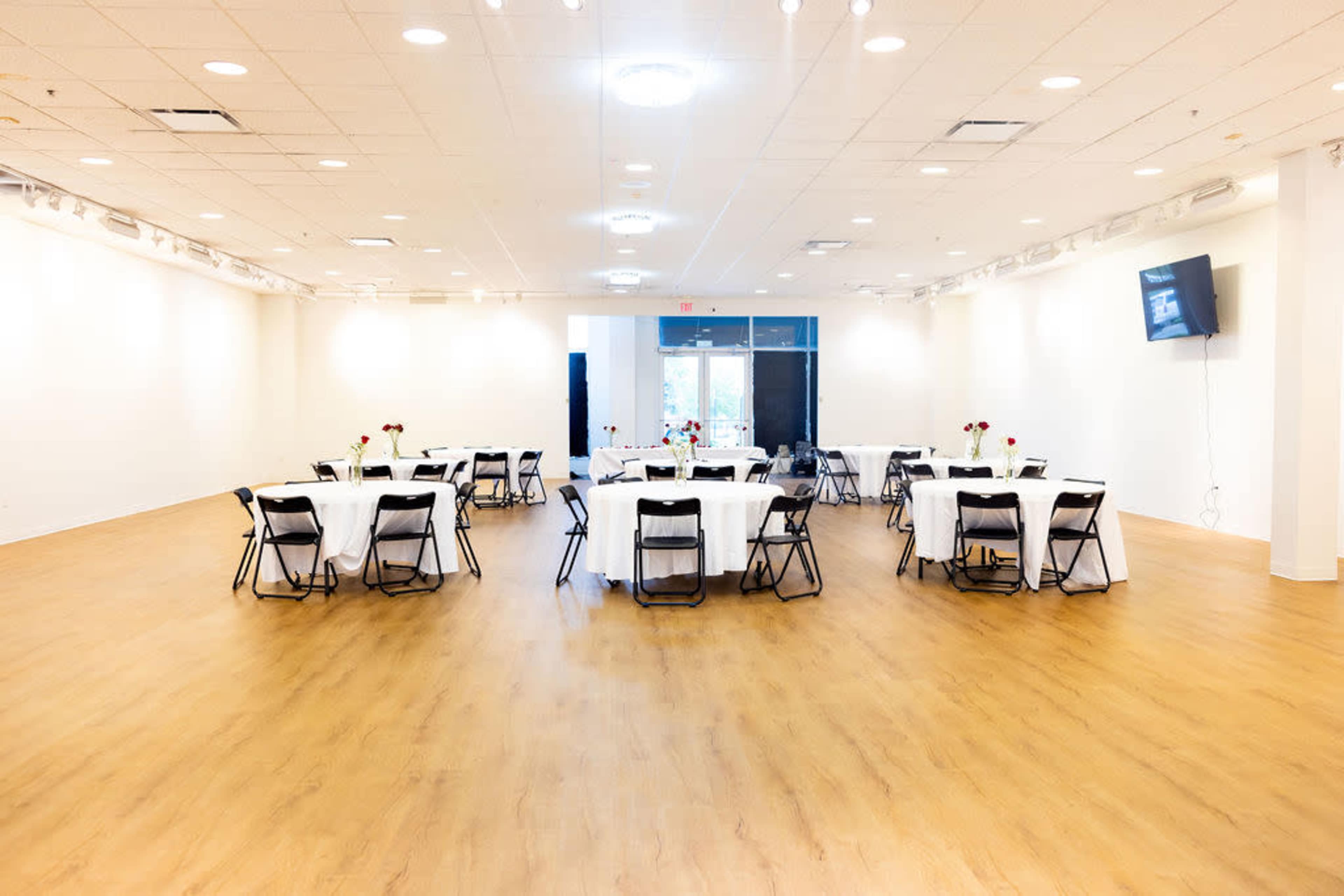 The image shows a spacious event room set with multiple round tables covered in white tablecloths and black chairs arranged around them, with a few roses as centerpieces.