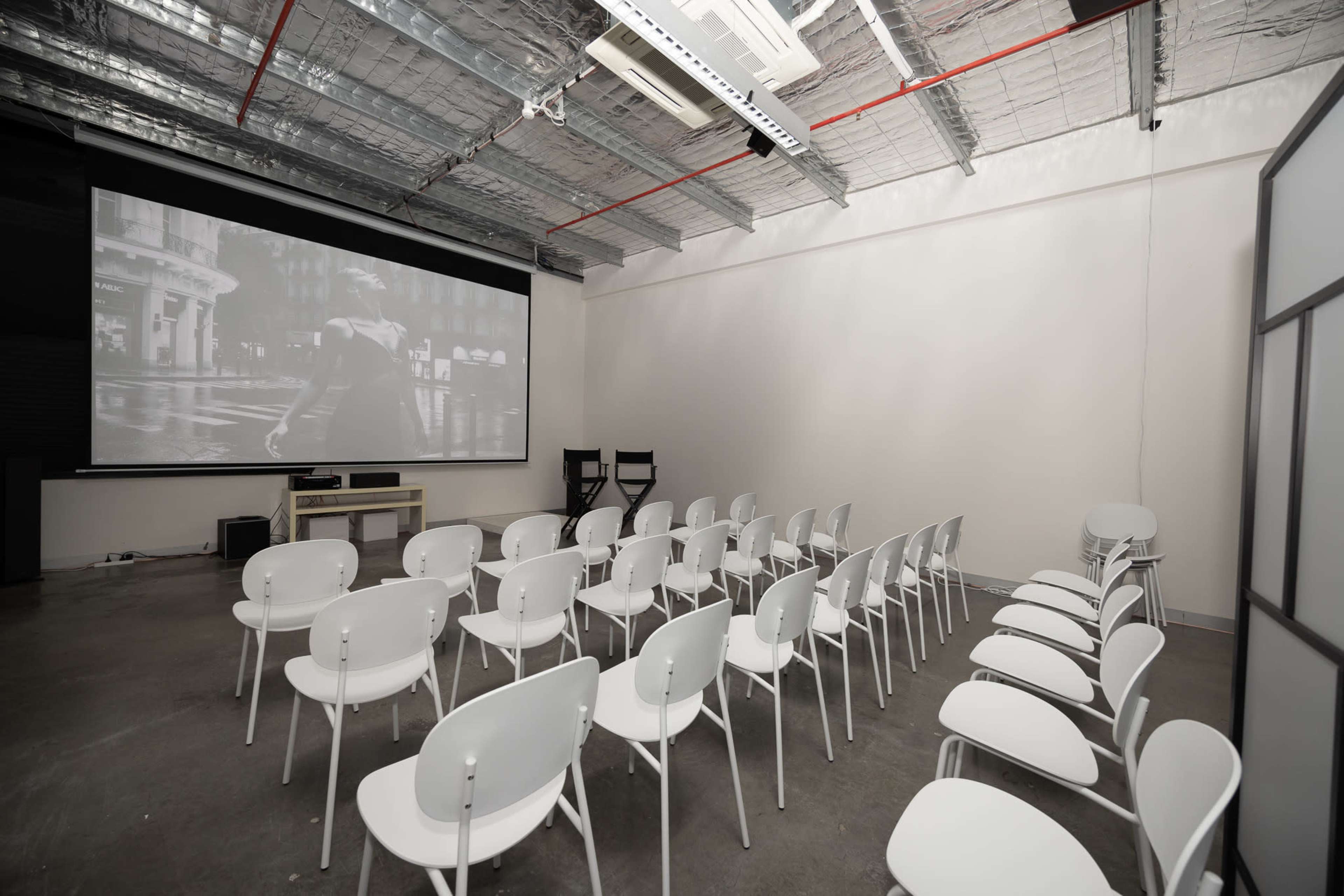 The image shows a sparsely filled room set up for an event, featuring rows of white chairs facing a large projection screen displaying a black-and-white video.
