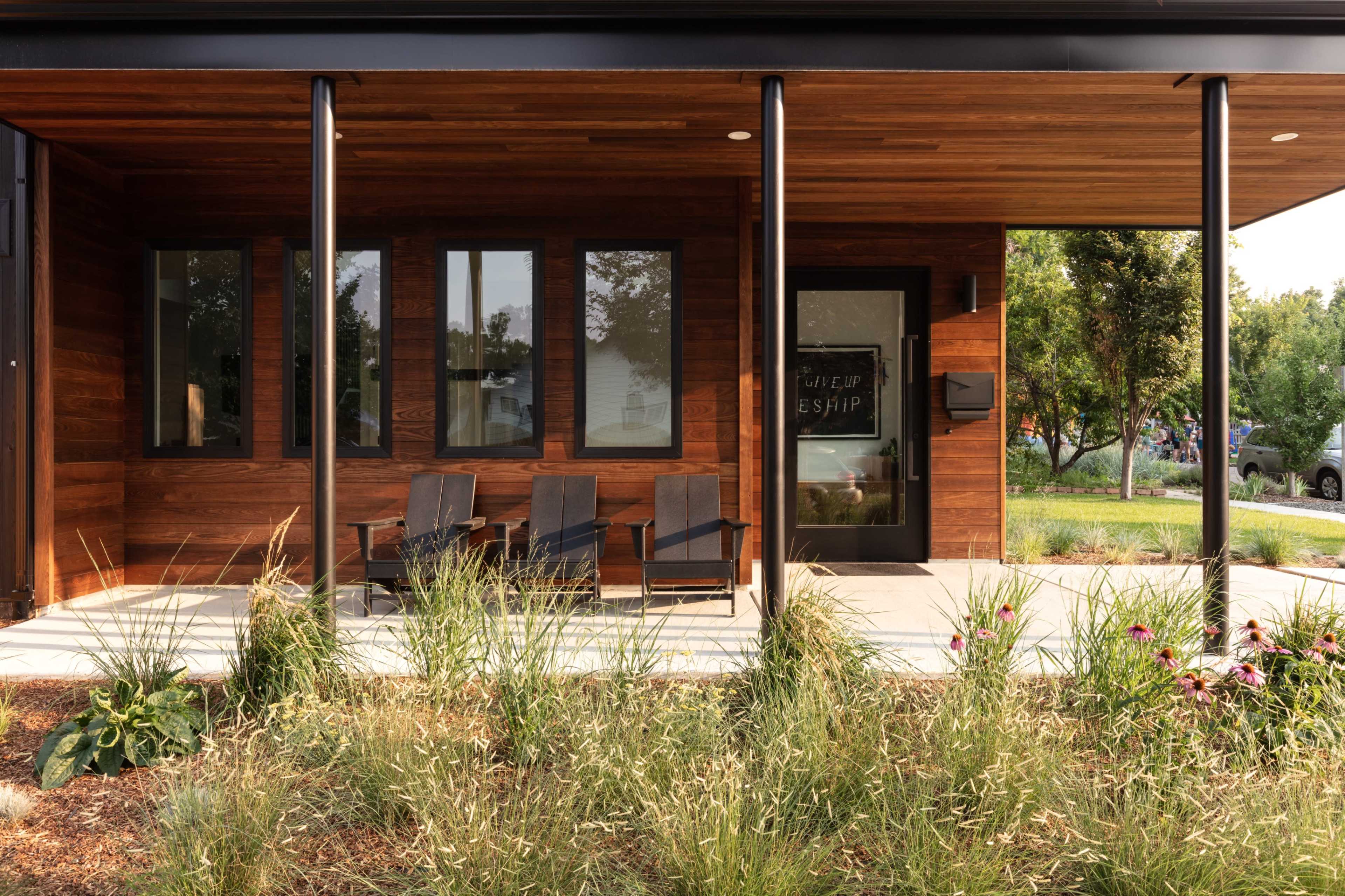 The image shows a modern house with a wooden facade, featuring a porch with three black Adirondack chairs and a landscaped yard.