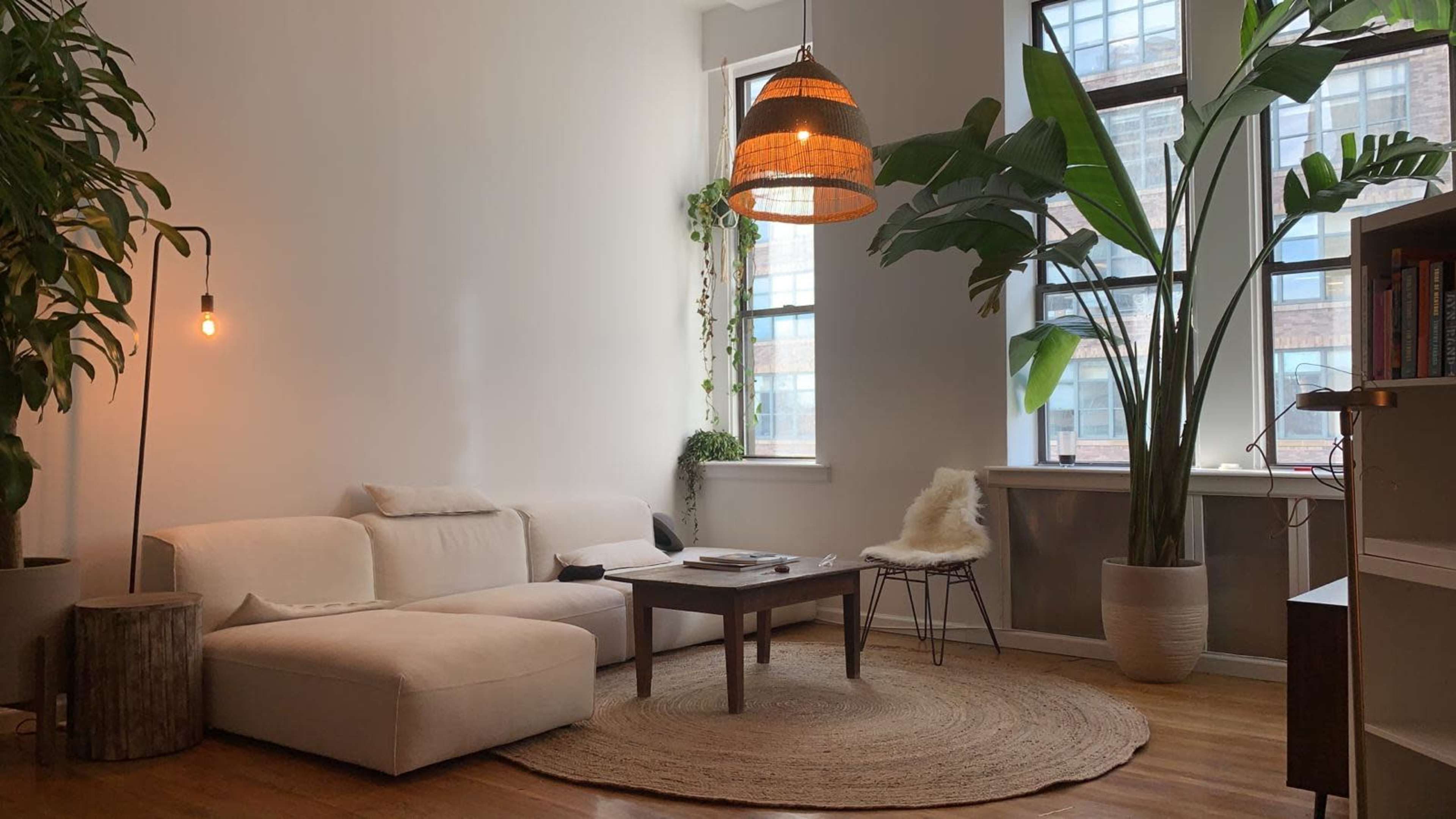 A spacious living room features a white sectional sofa, a round coffee table, and large windows with indoor plants.