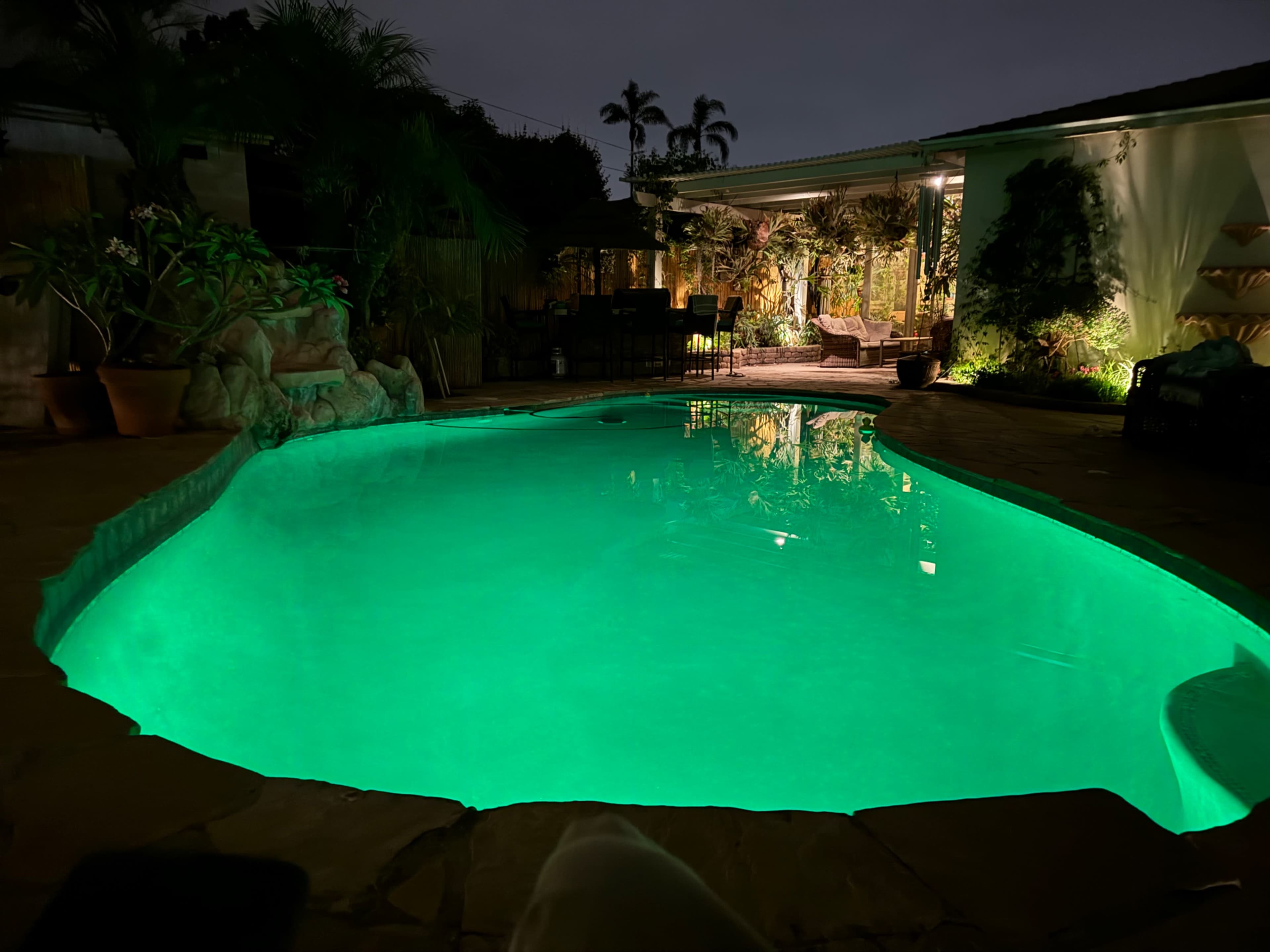 The image shows a illuminated swimming pool surrounded by lush plants and a patio area at night.