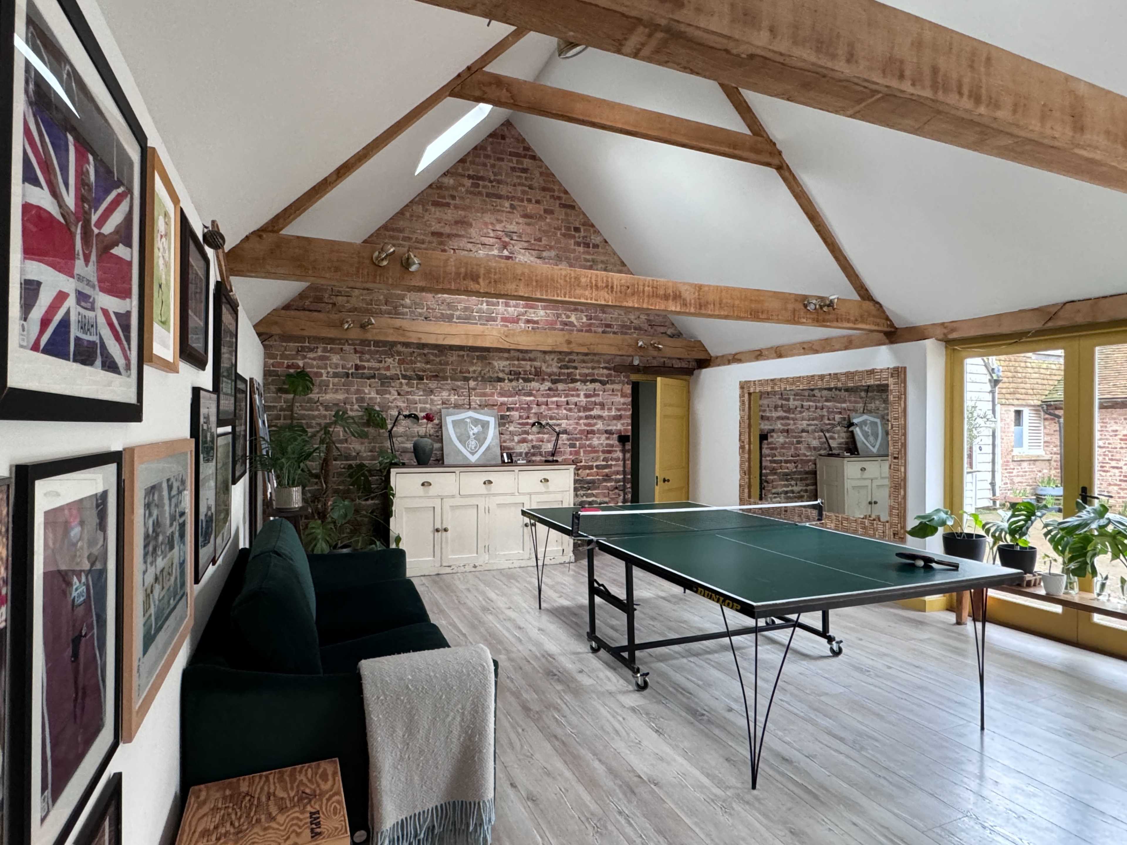 A spacious game room with a table tennis table, exposed brick walls, wooden beams, and multiple potted plants.