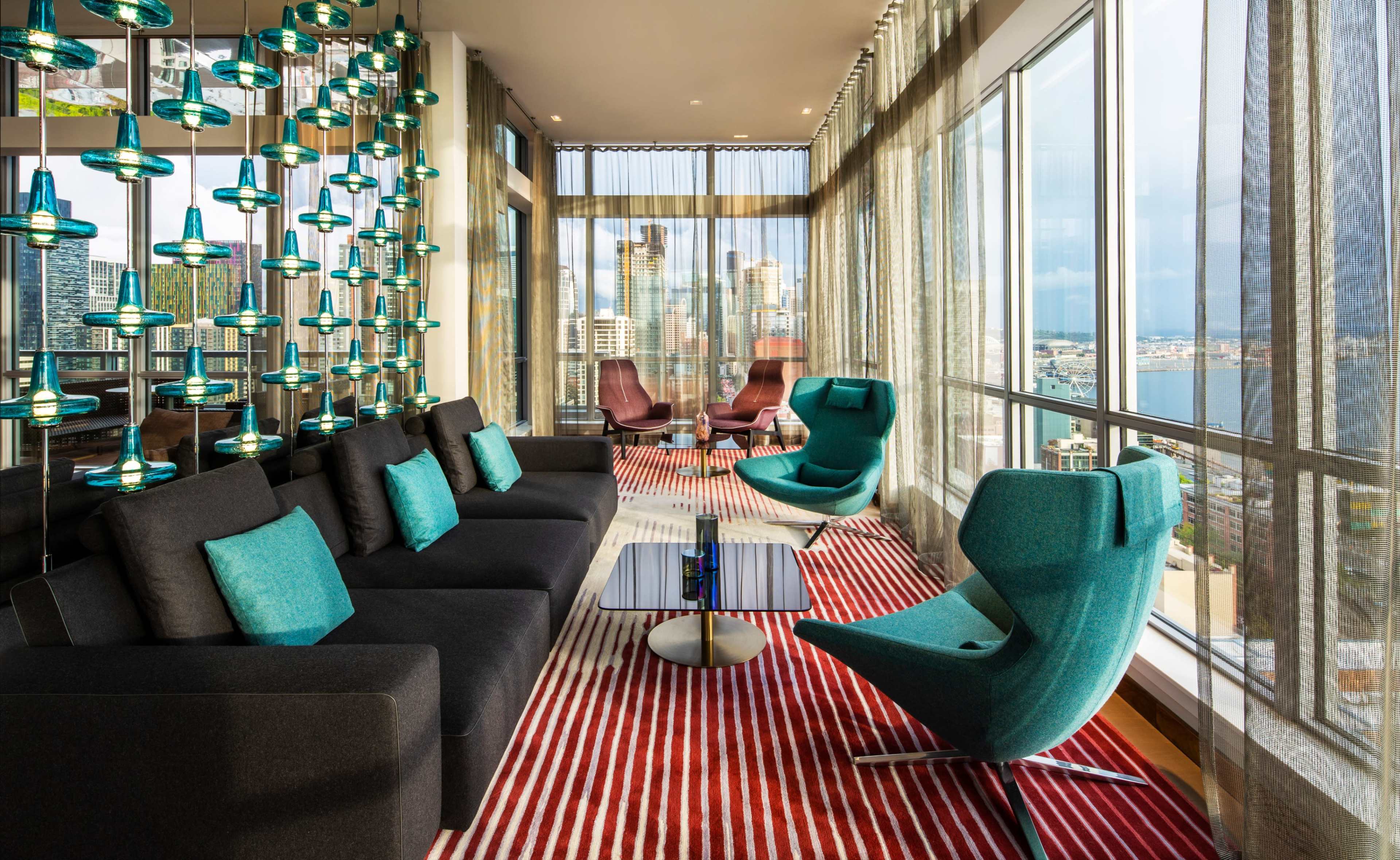 A modern lounge features a sleek sofa and chairs with turquoise accents, large windows offering a city view, and decorative lighting.