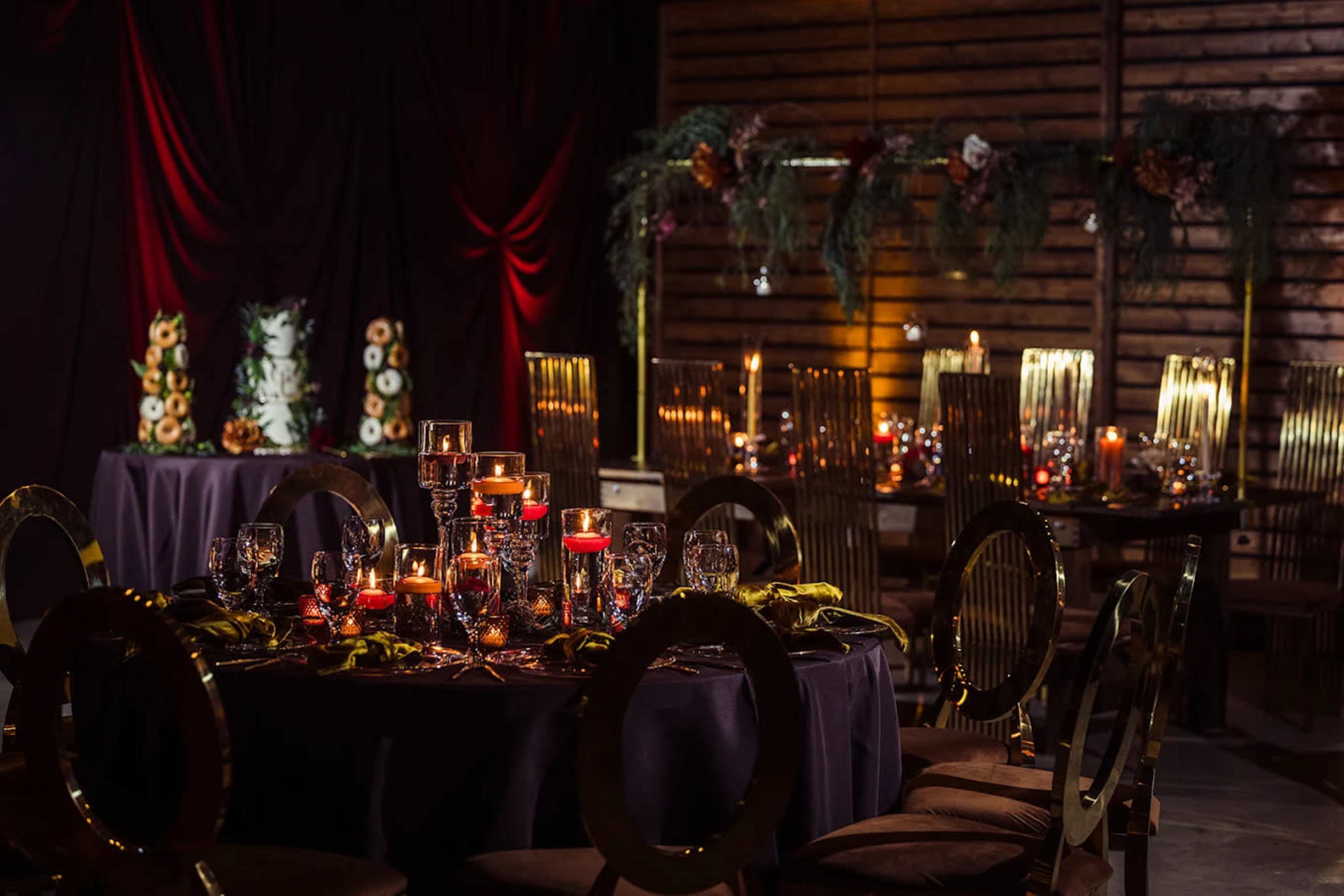 The image shows a decorated dining area with elegantly set tables featuring candles, glassware, and a backdrop of wooden panels and draped fabric.