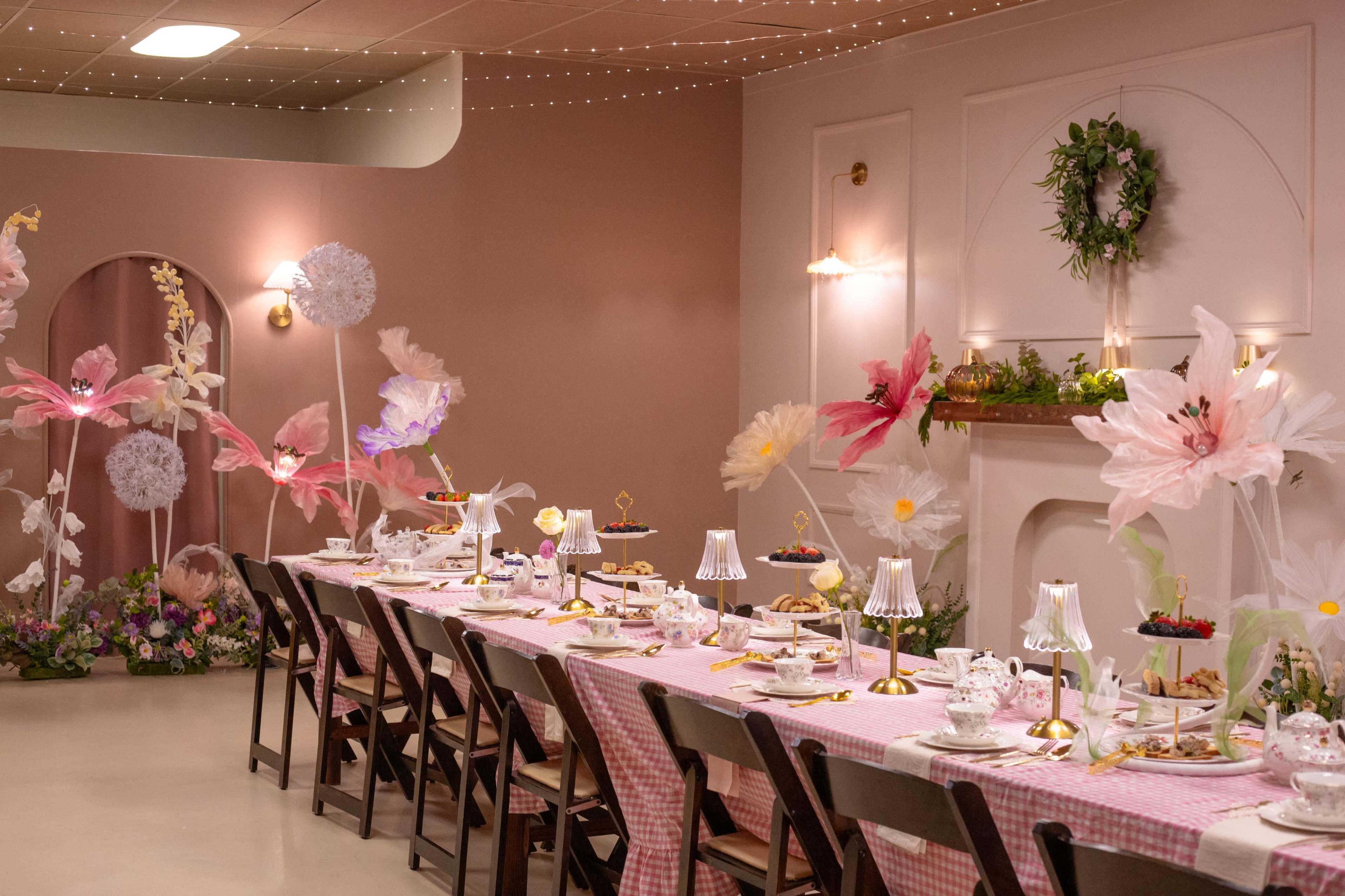 The scene features a long dining table set with floral decorations and elegant tableware in a softly lit room adorned with pink walls and whimsical paper flowers.