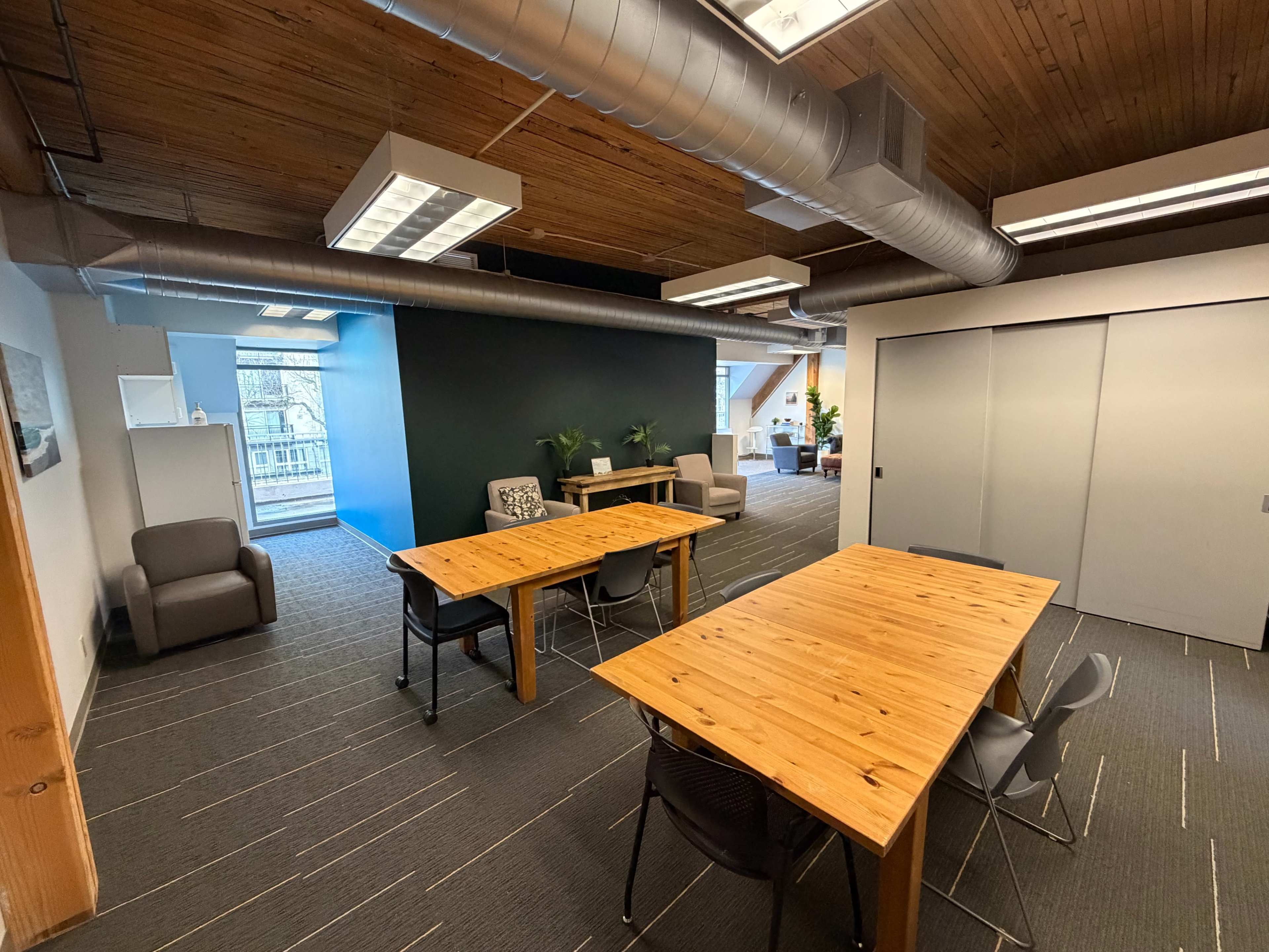The image shows an office space featuring two wooden tables surrounded by chairs, with a blue accent wall and large windows providing natural light.