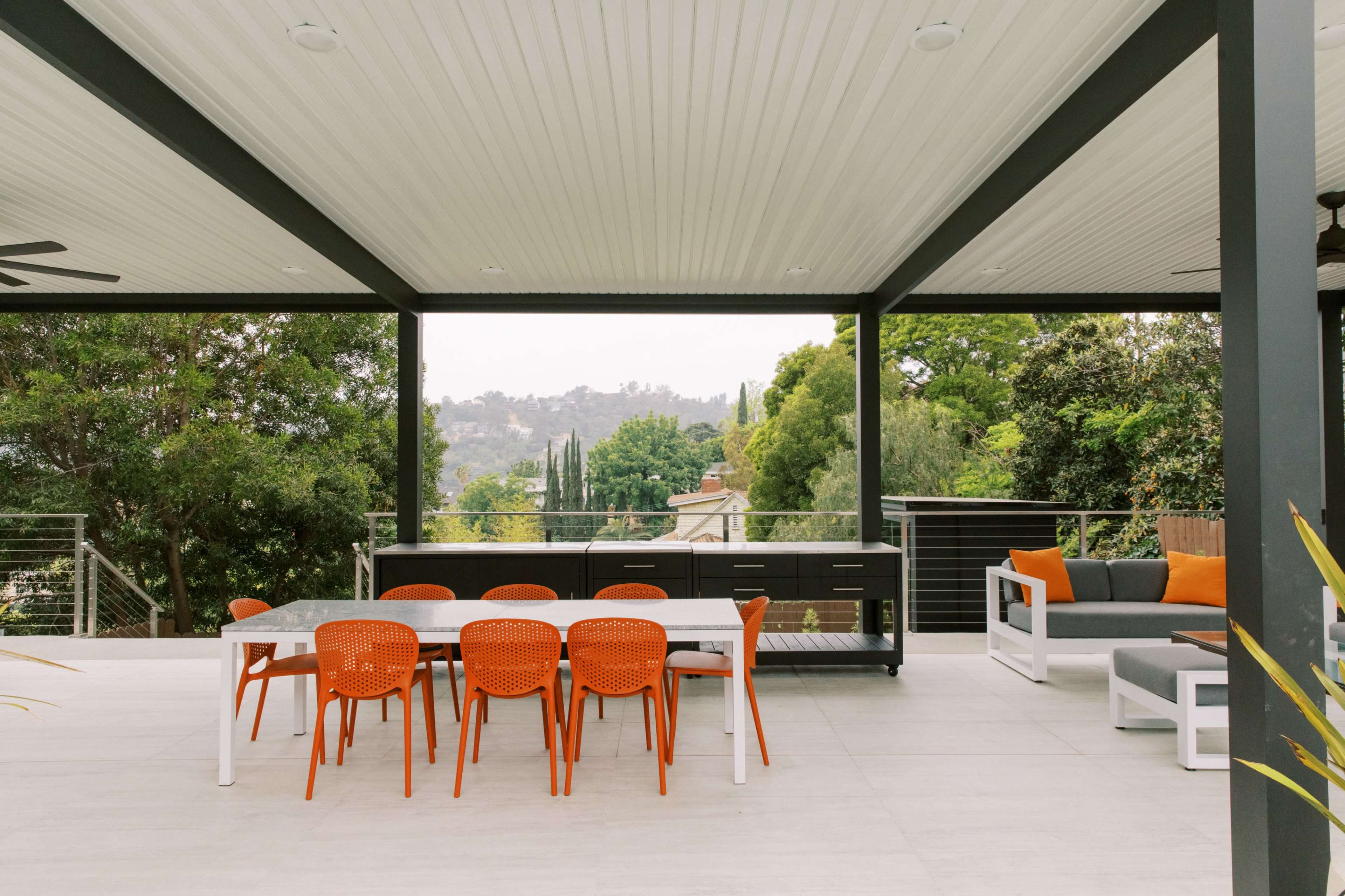 The image shows an outdoor patio area featuring a dining table with orange chairs, a sectional sofa with orange cushions, and a view of greenery in the background.