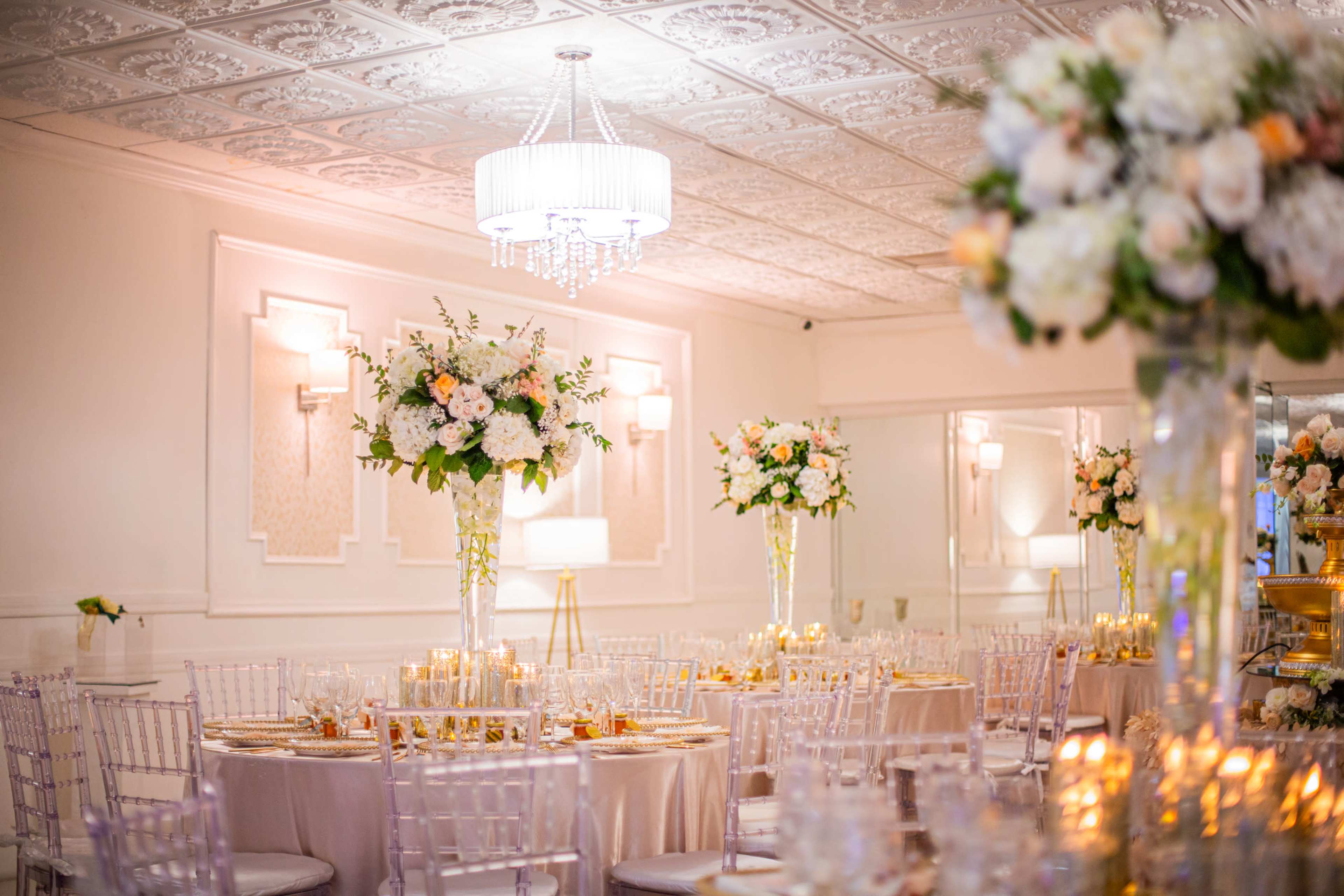 The image shows an elegantly arranged banquet hall with round tables set with floral centerpieces, soft lighting, and a decorative ceiling.