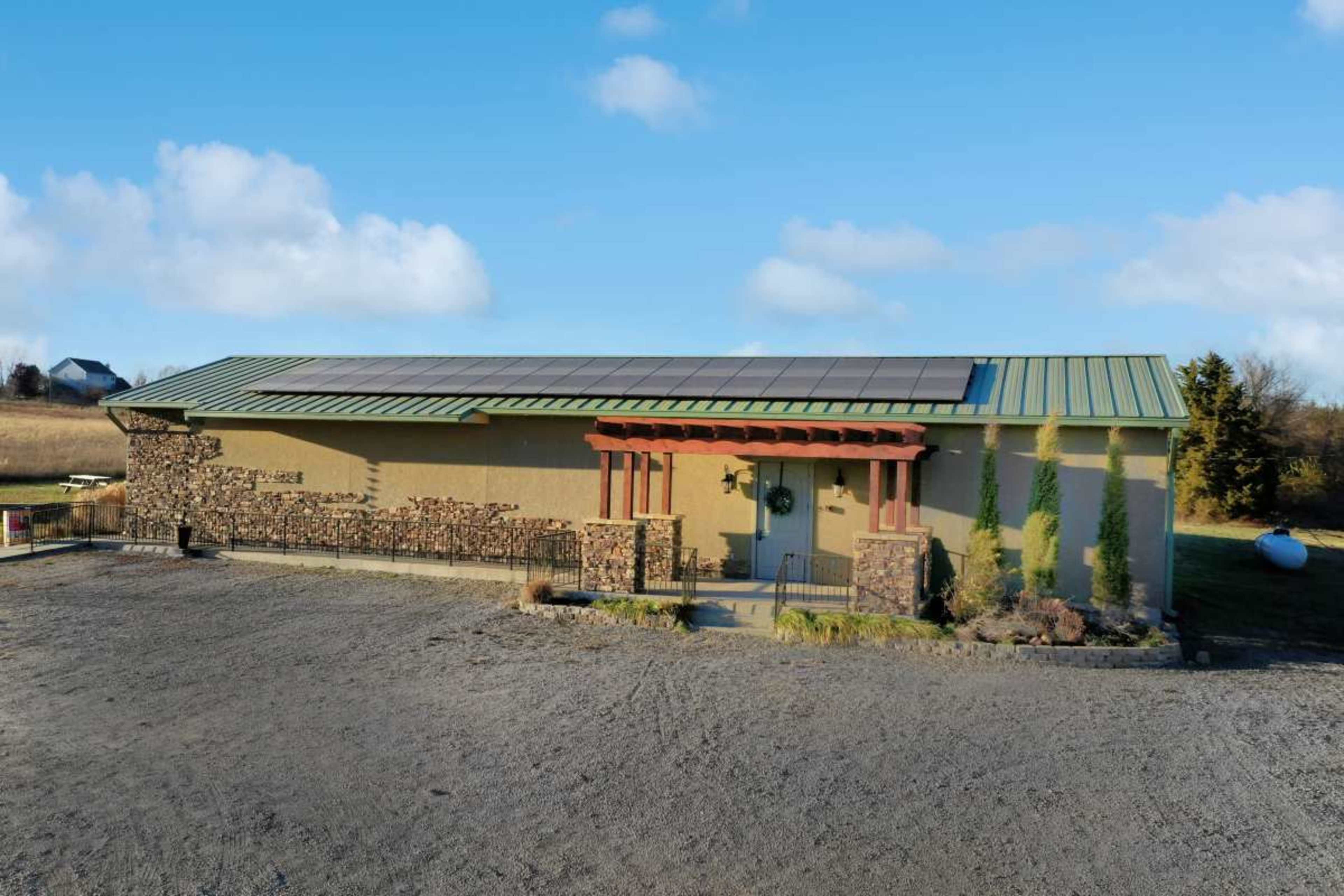 The image shows a single-story building with a stone facade, a green metal roof, and solar panels on the roof, situated in an open area.