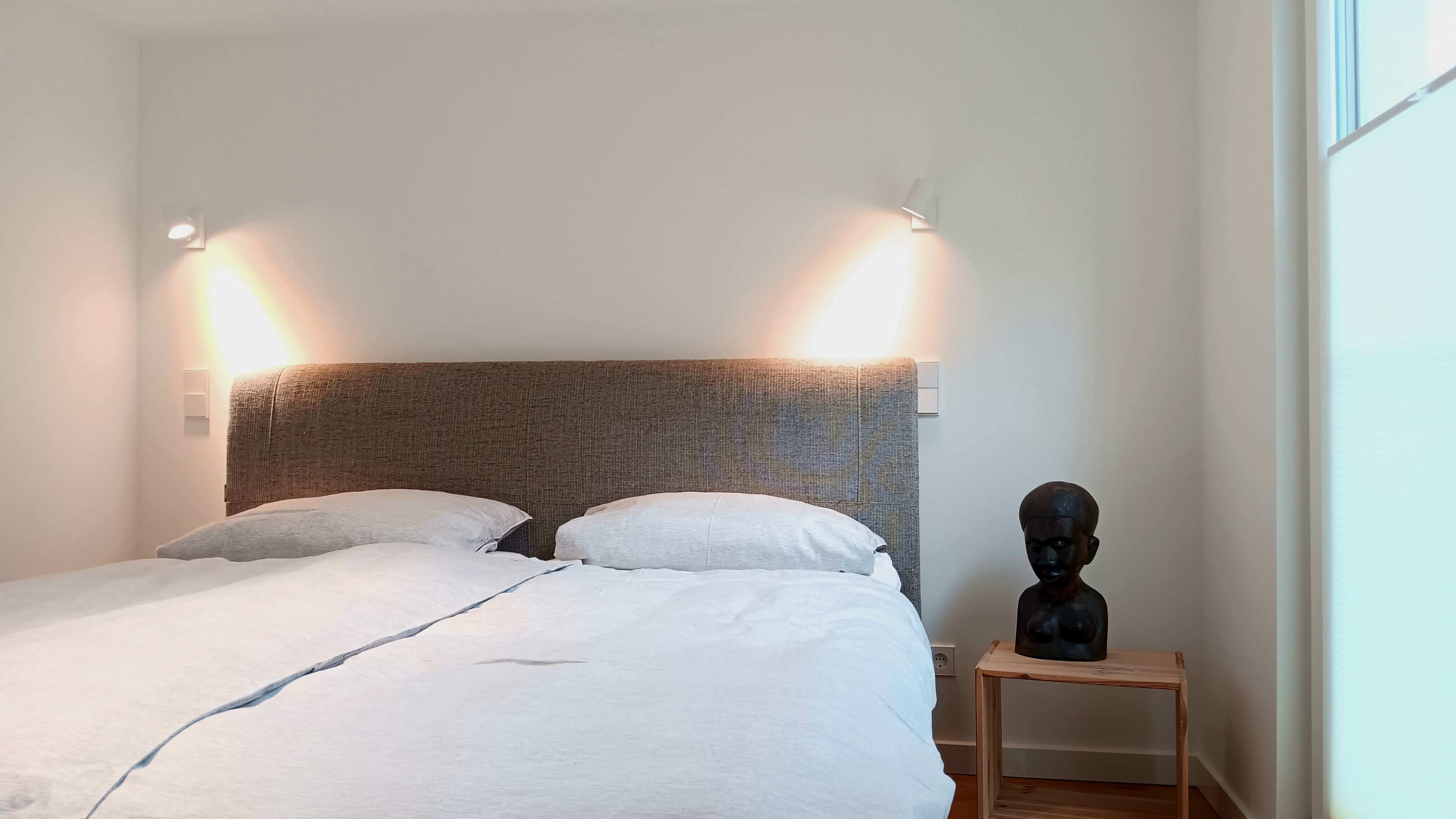 A neatly made bed with white linens is positioned against a wall, accompanied by a bedside table featuring a dark sculpture.