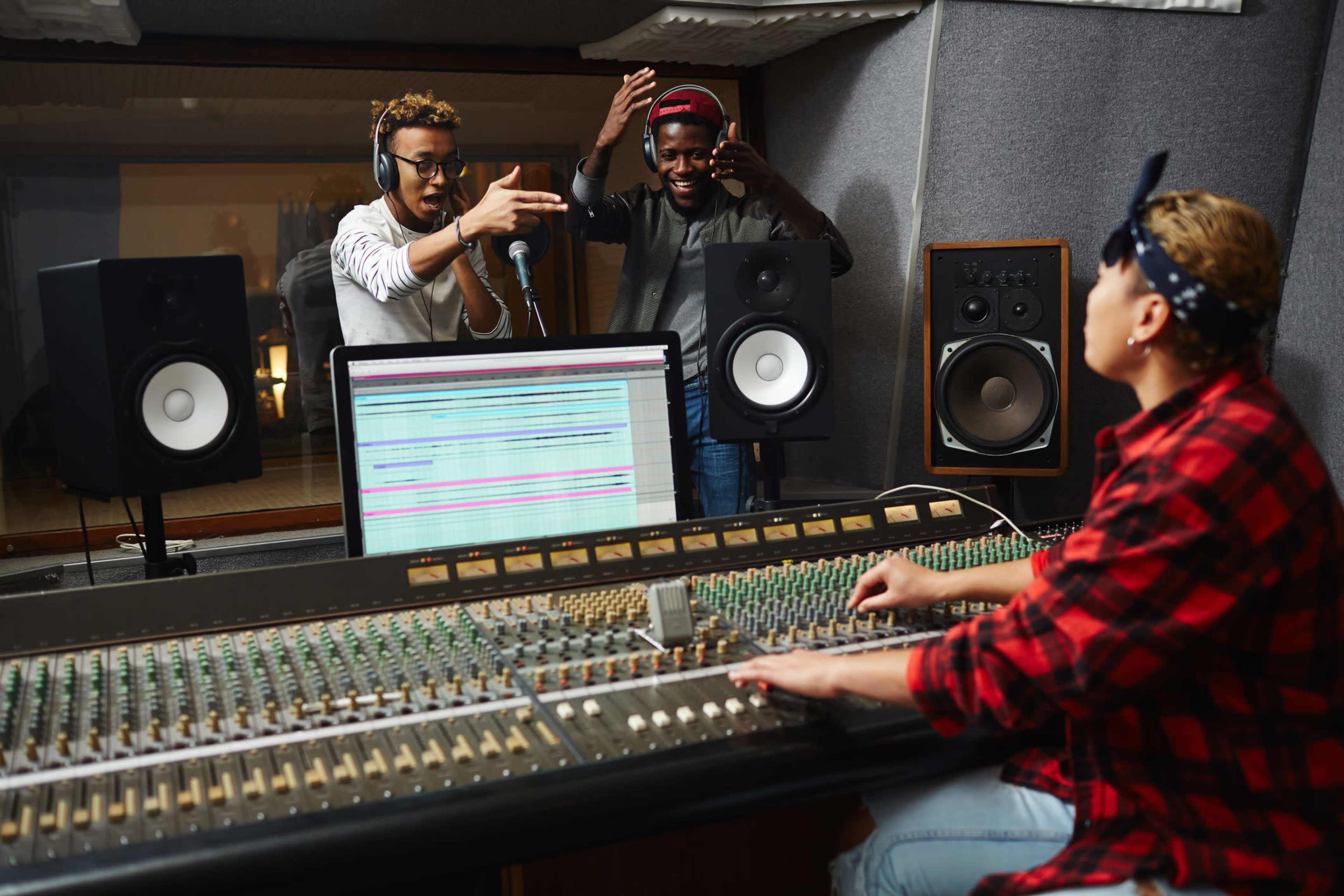 Two recording artists perform in a sound booth while a sound engineer operates the mixing console in a music studio.