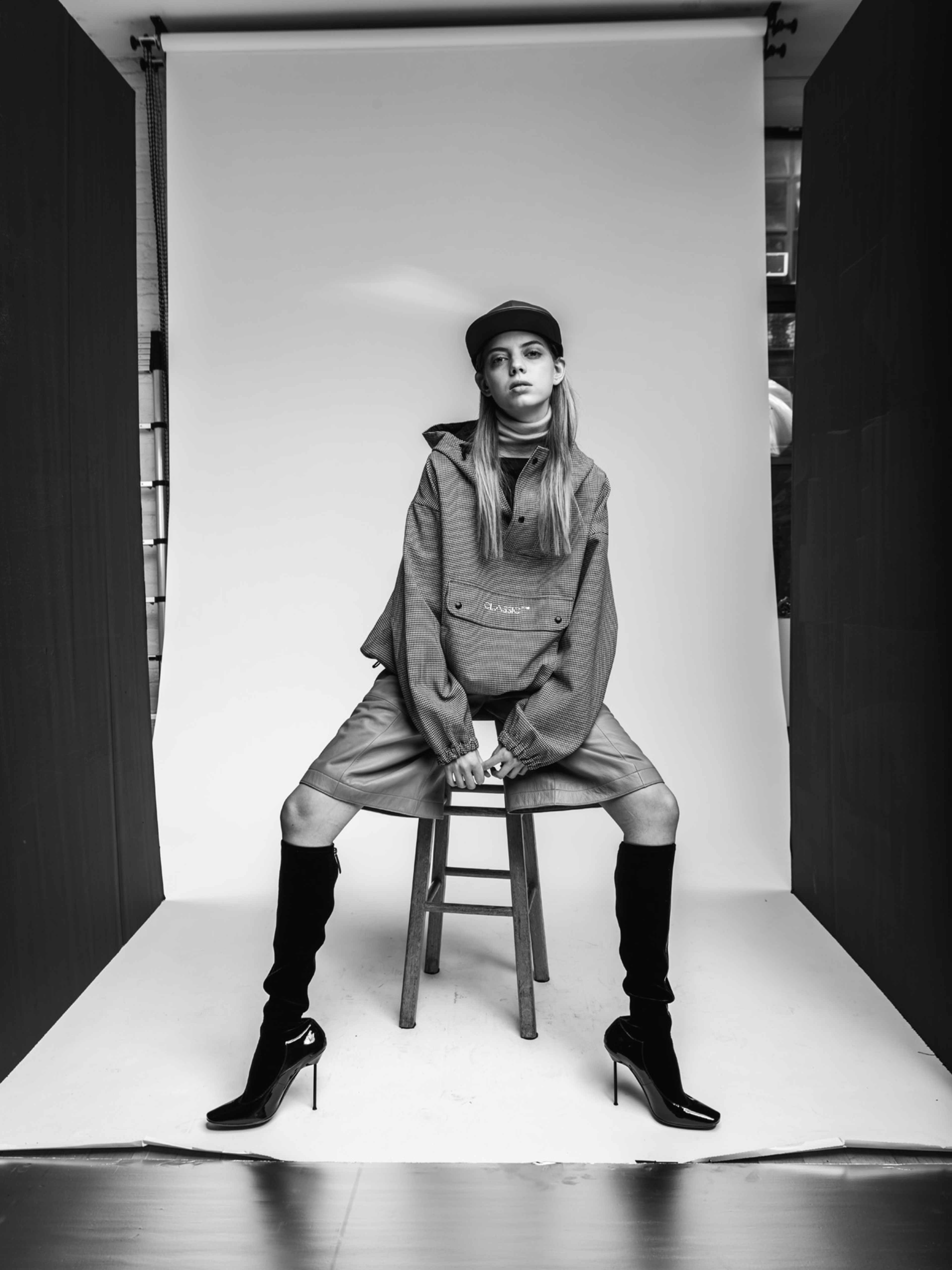 A model in stylish attire sits on a stool against a plain backdrop in a black-and-white photo.