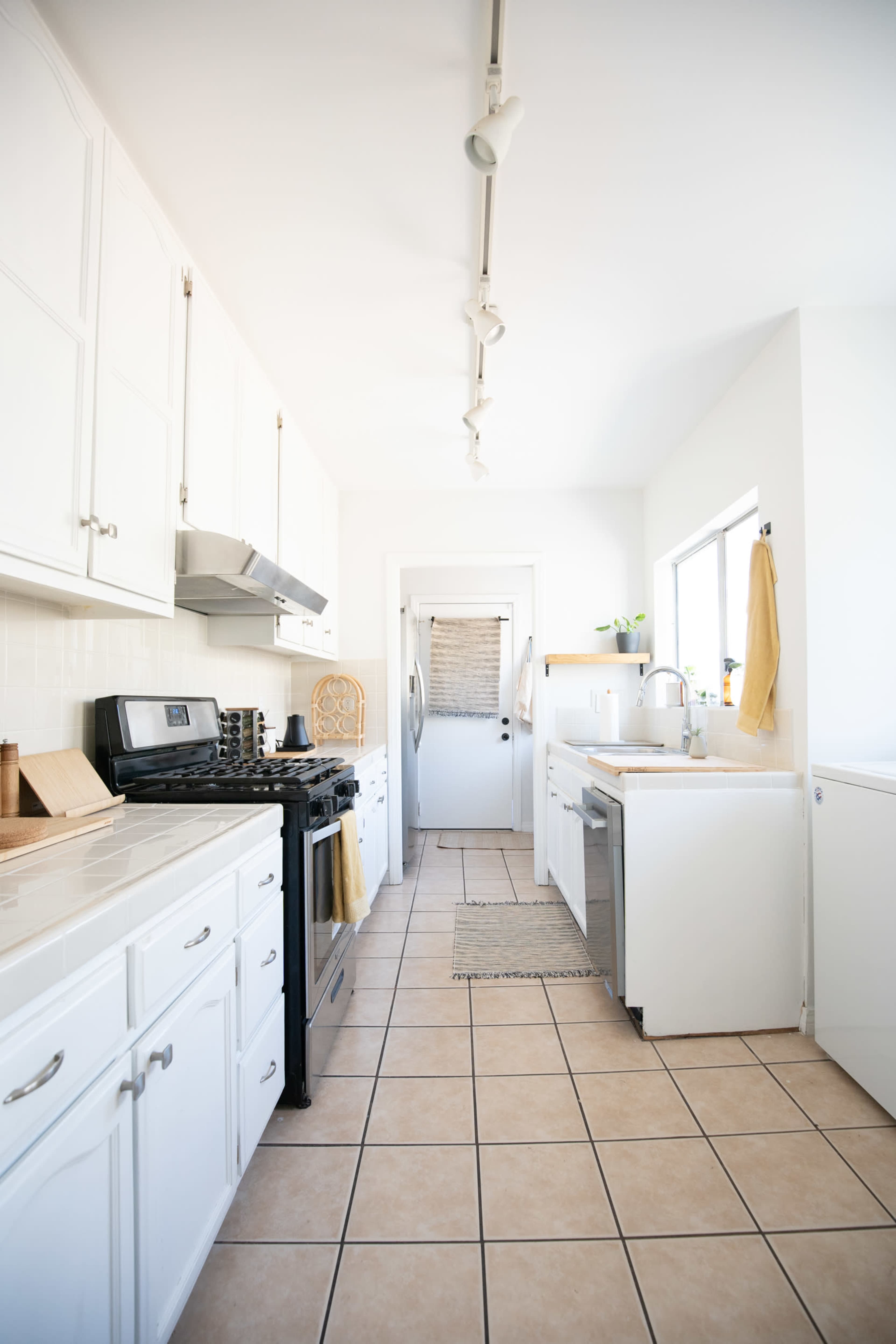 A long, narrow kitchen with white cabinetry, a gas stove, stainless steel appliances, and beige tiled flooring.