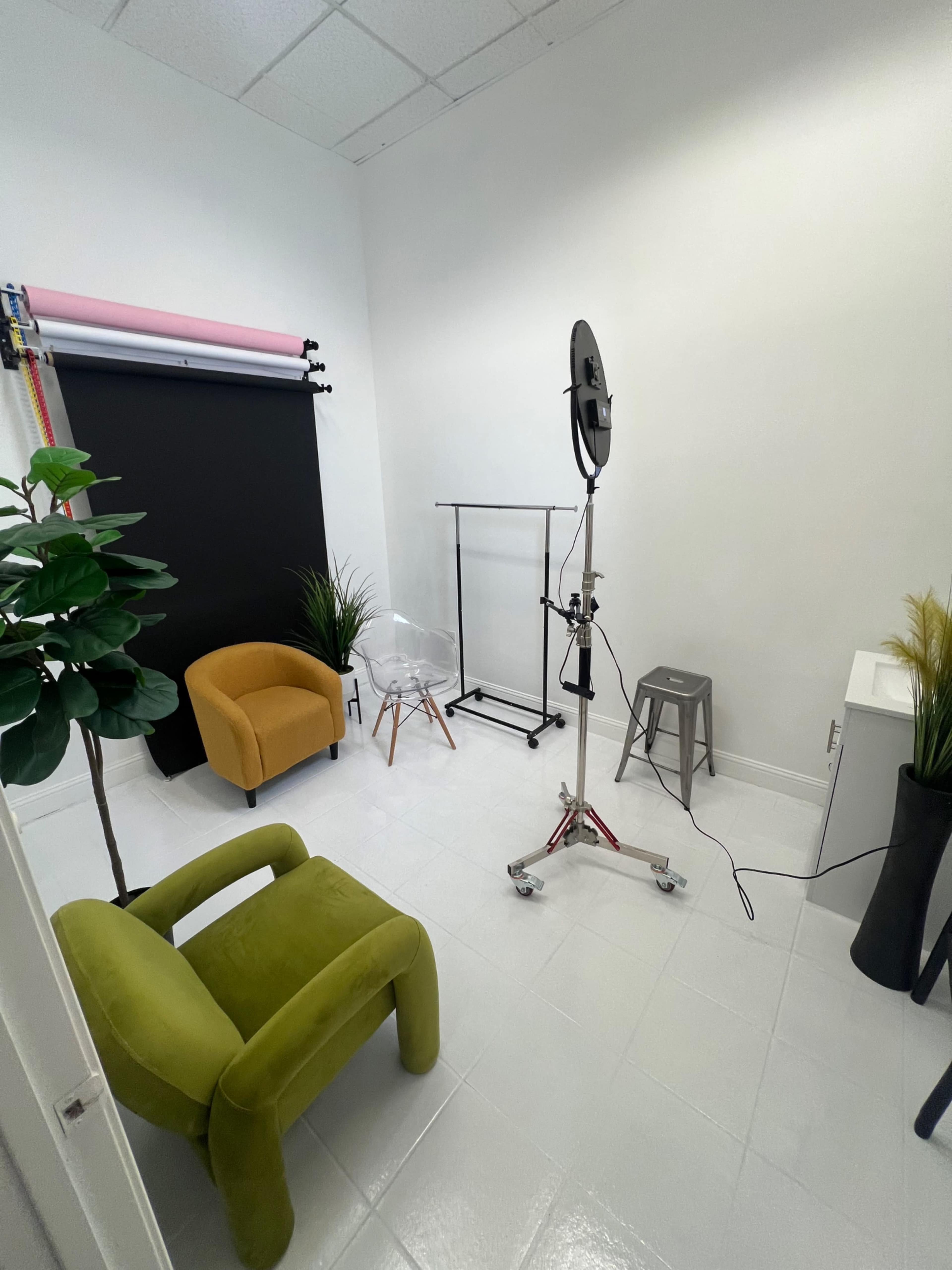 The image shows a small photography studio featuring a green armchair, a yellow armchair, a backdrop stand ready for setup, a clothing rack, and various decorative plants.