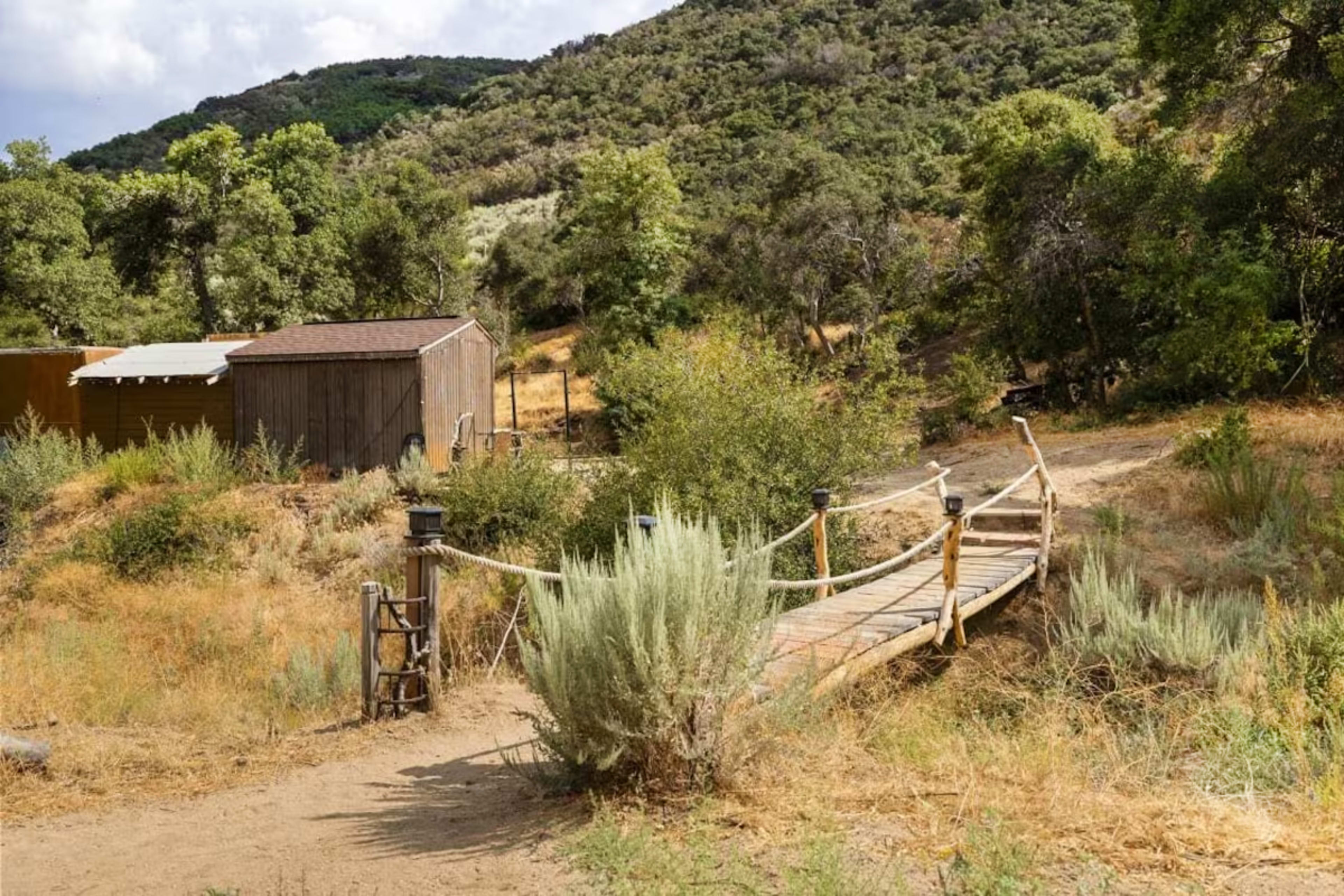 A wooden bridge leads across a dry area to a small shed surrounded by shrubs and trees on a hillside.