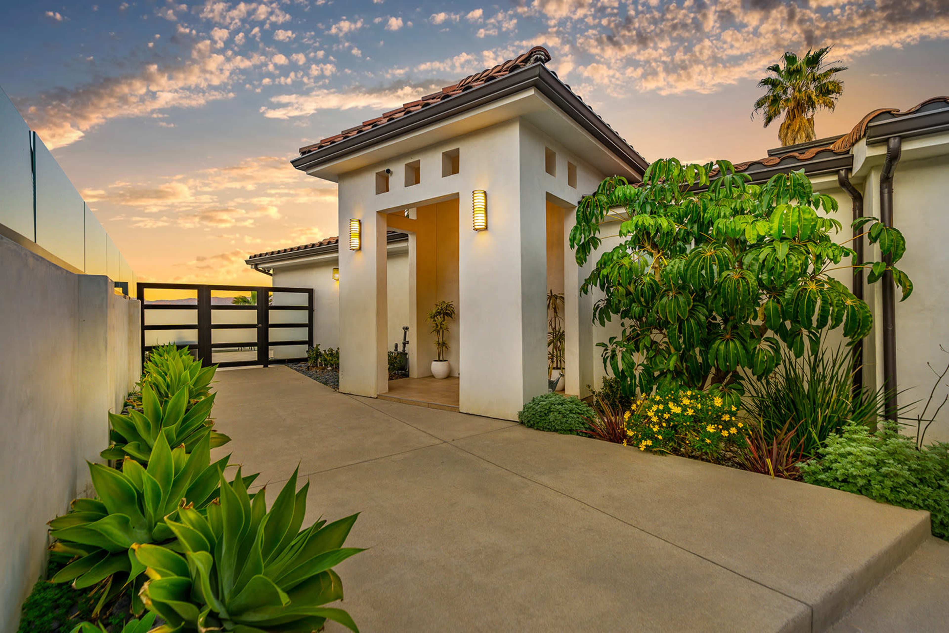A modern entrance to a home, featuring a walkway framed by plants and a decorative gate under a sunset sky.