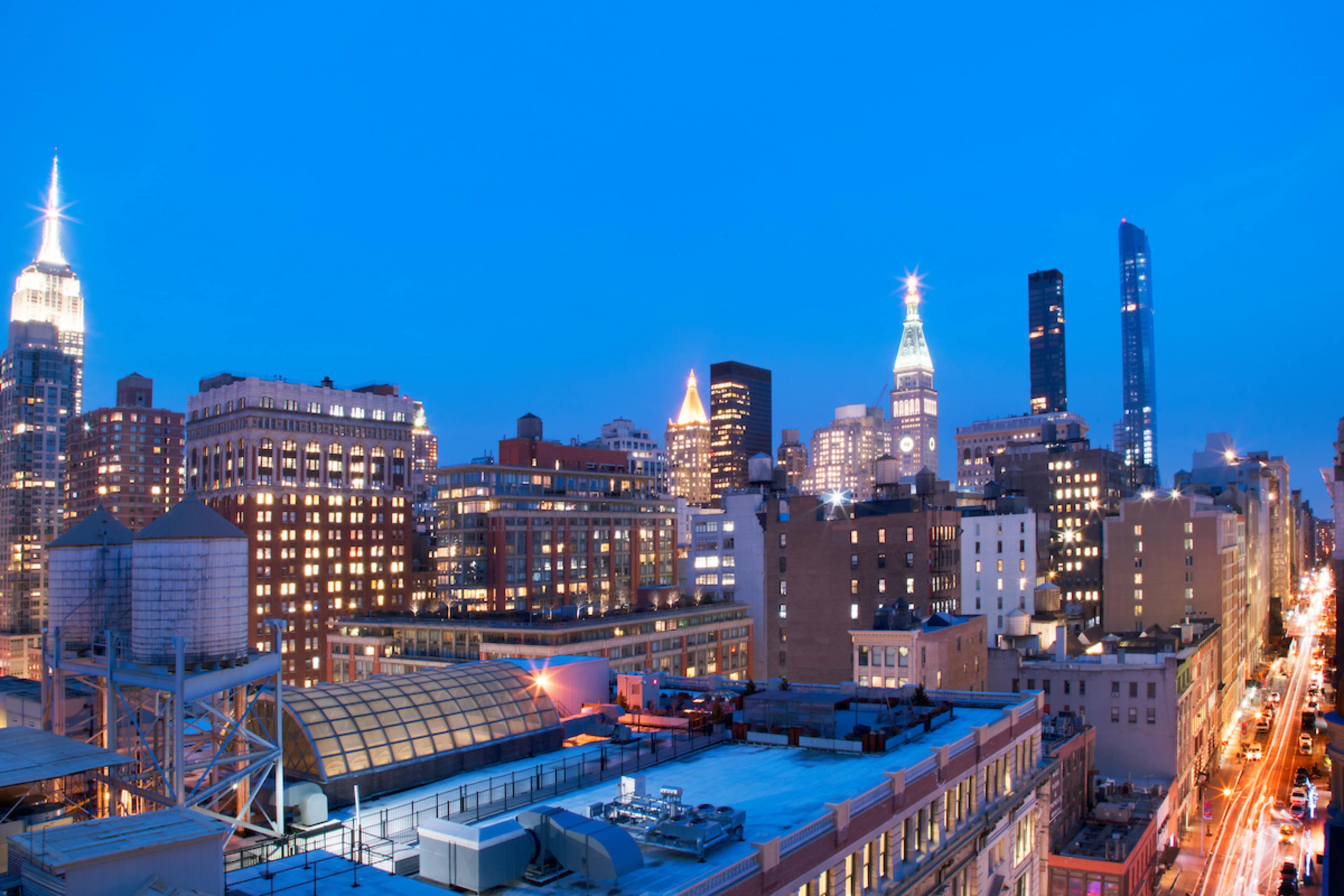 The image shows a skyline of New York City at twilight, featuring illuminated buildings including the Empire State Building and other skyscrapers along a bustling street.