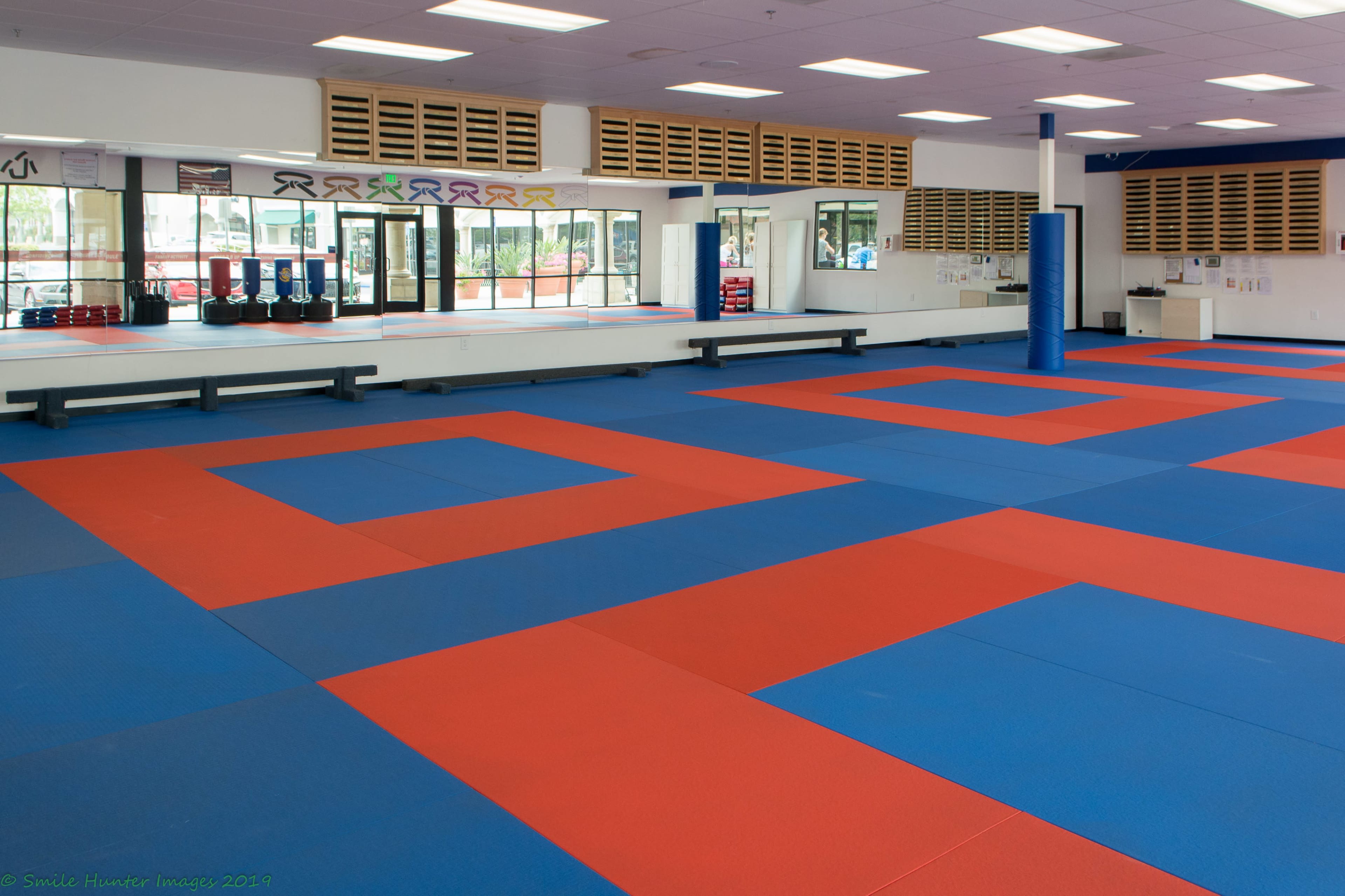 A martial arts training facility with blue and red mats covering the floor, and large mirrors along one wall.