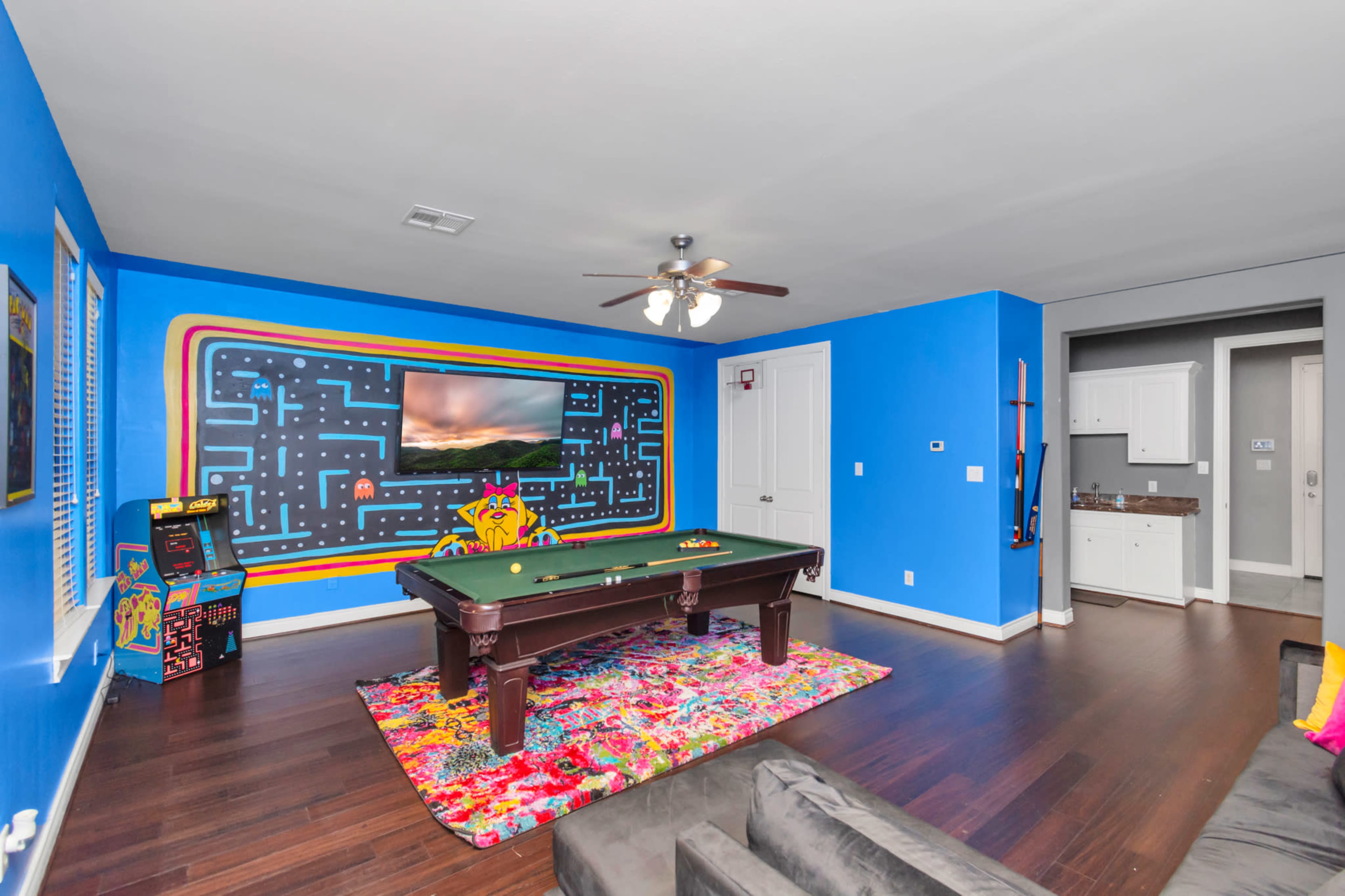 The room features a bright blue wall with a Pac-Man mural, a pool table in the center, and an arcade machine in the corner.