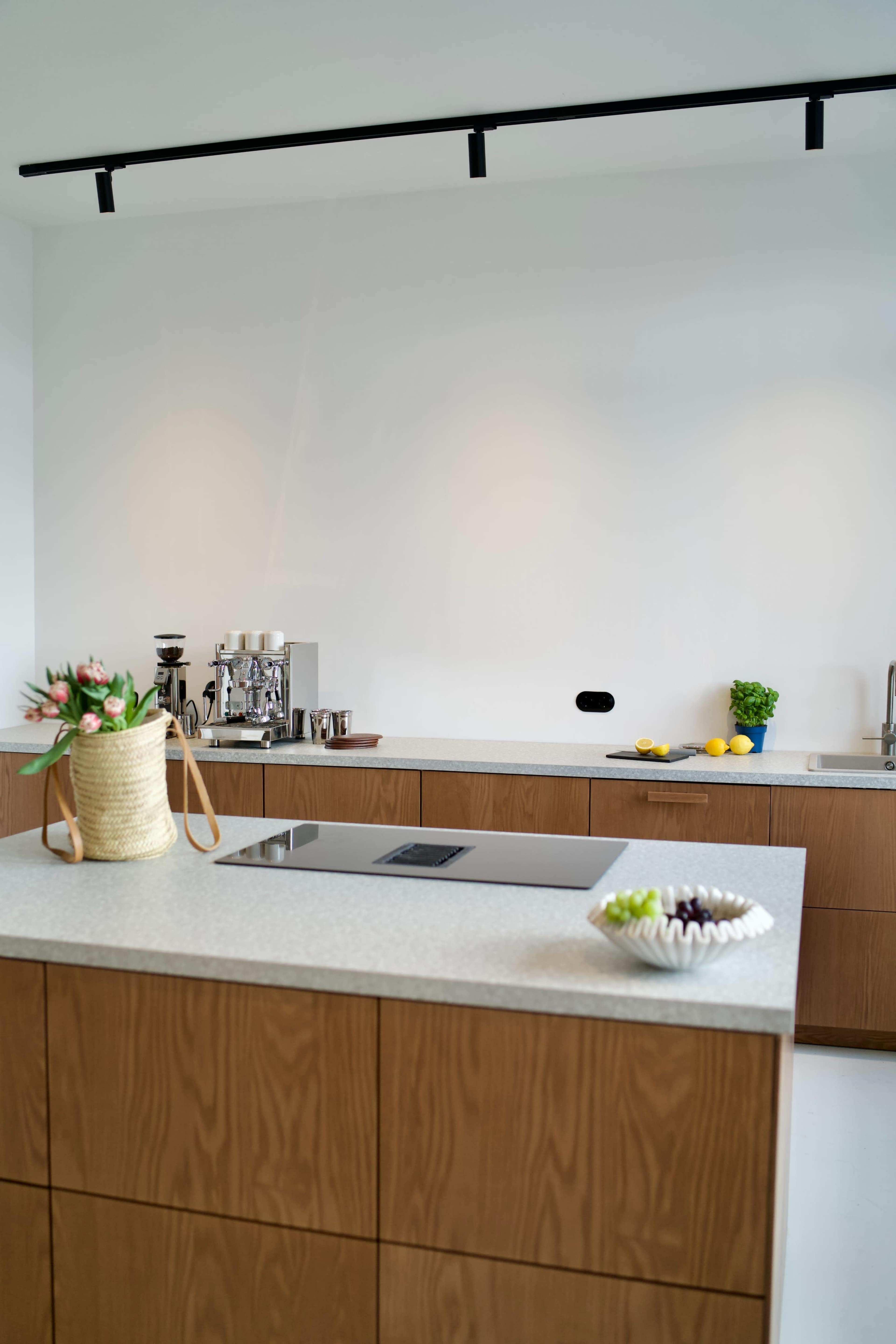 The image depicts a modern kitchen featuring a central island with a cooktop, wooden cabinetry, and a countertop adorned with fruits and a vase of flowers.