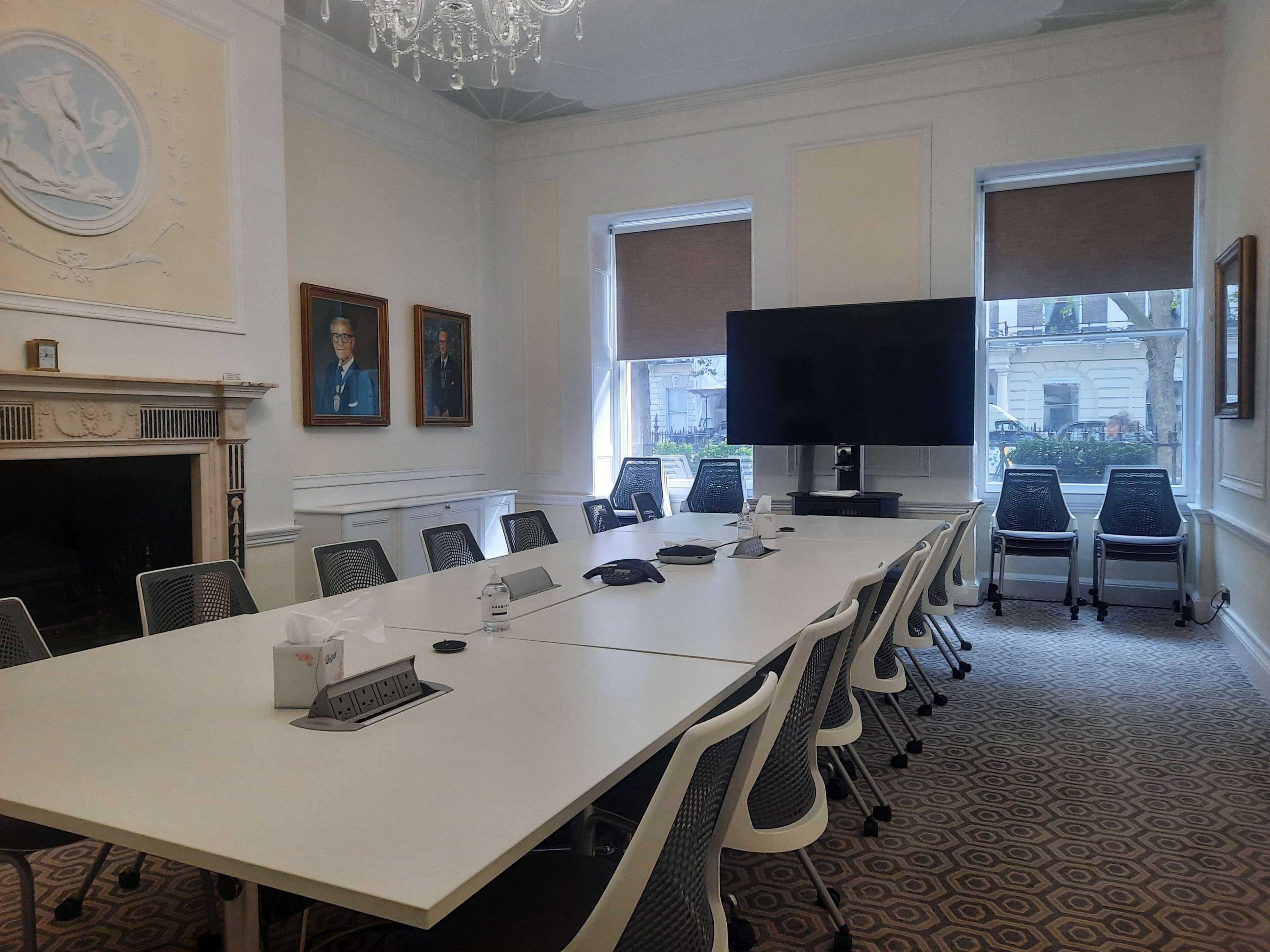 A conference room with a long white table, black chairs, a large screen on one wall, and framed portraits on the adjacent walls.