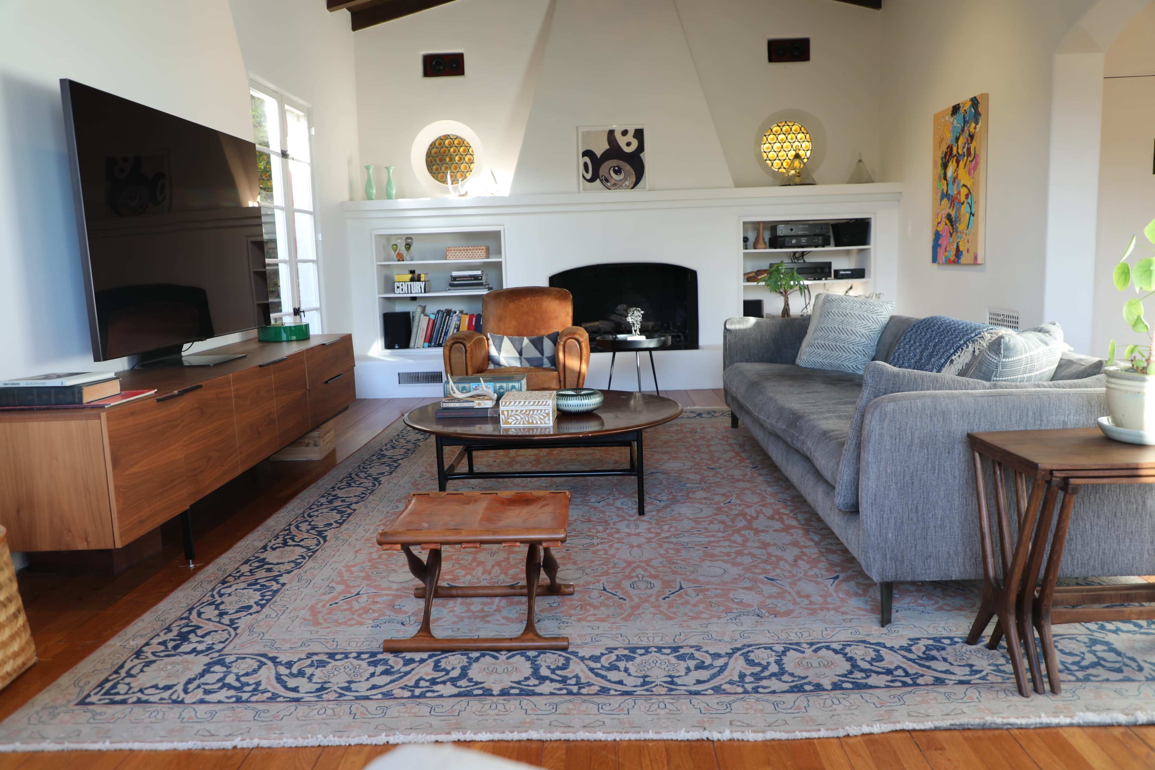 The living room features a gray sofa, a wooden coffee table, a mid-century armchair, and a large television mounted on a wall unit, with decorative elements and artwork on display.