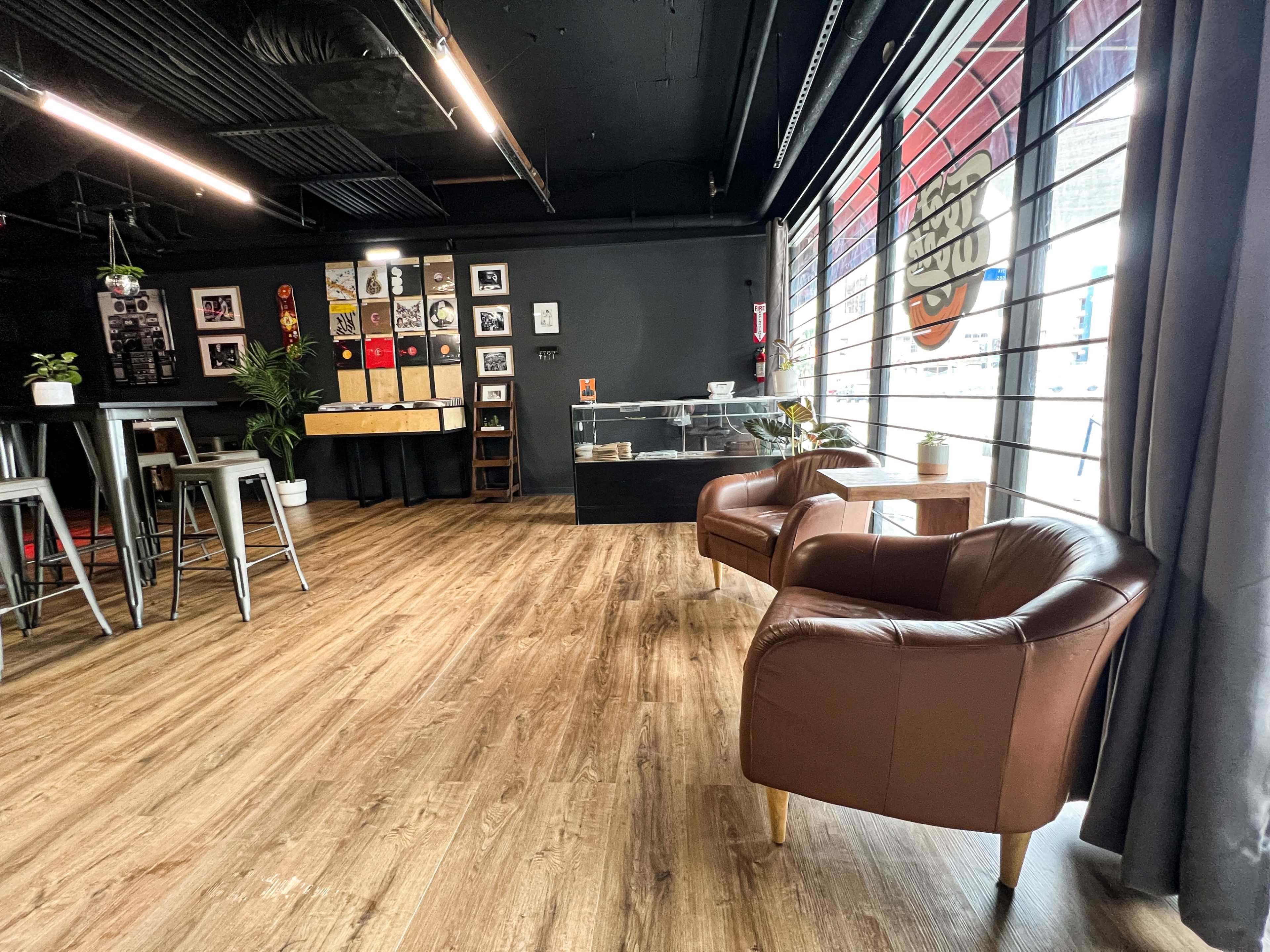 A modern café interior with wooden flooring, a display case, framed wall art, and two brown leather chairs facing a large window.