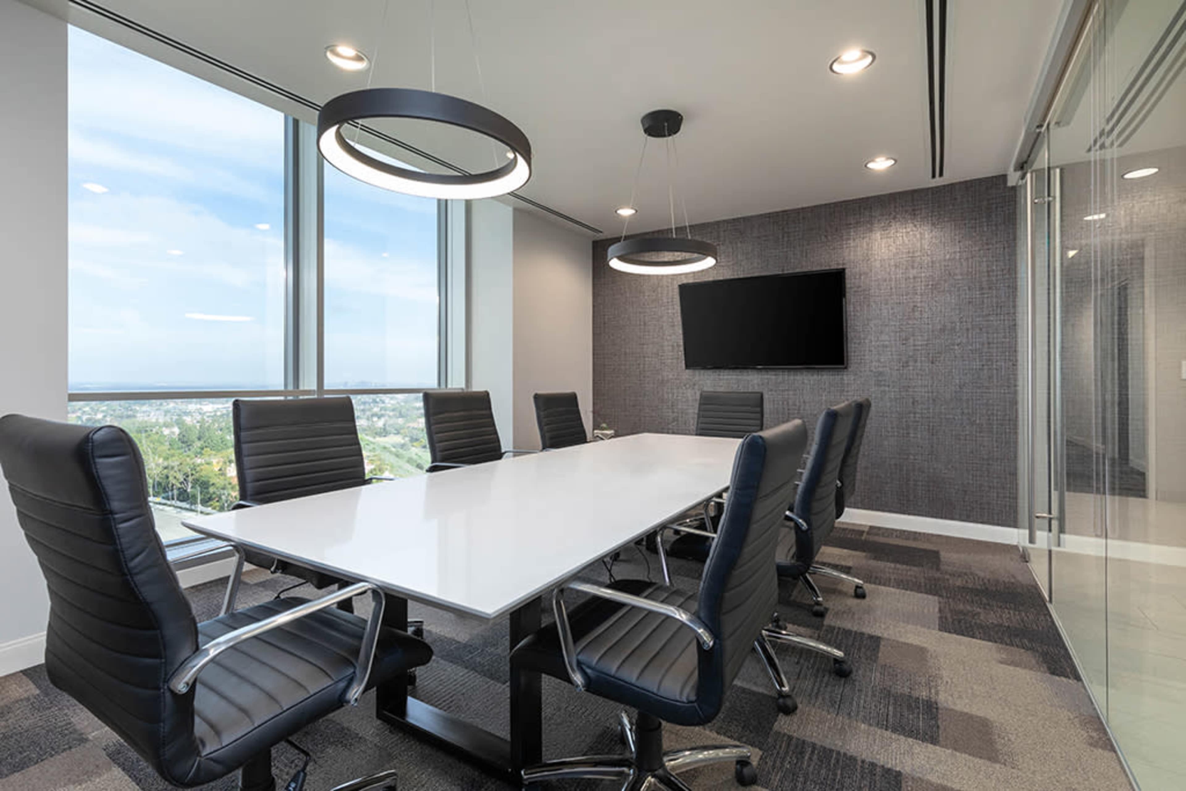 A modern conference room features a long white table surrounded by black leather chairs, with large windows offering a view and a wall-mounted TV.