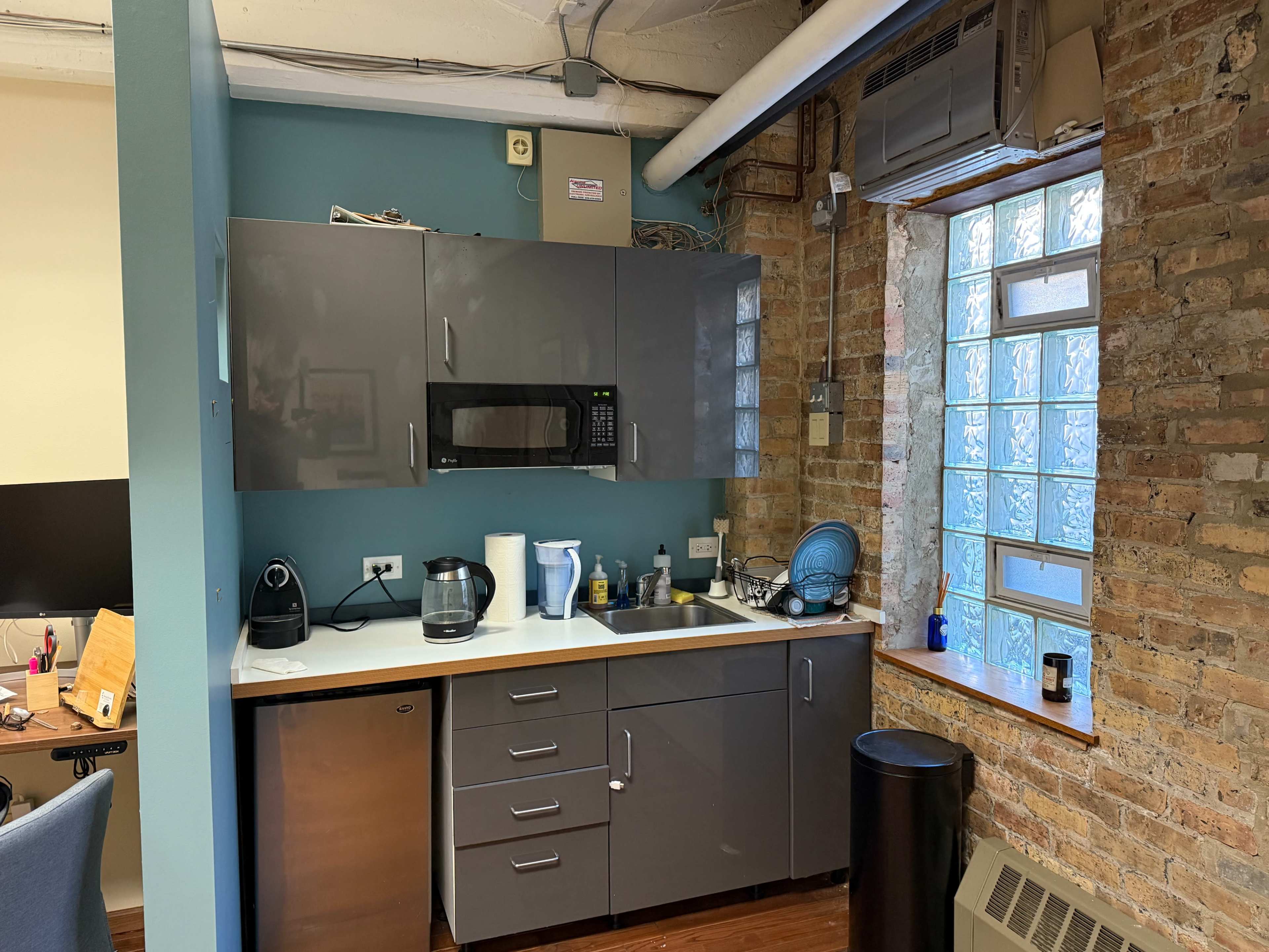 The image shows a modern kitchen area with gray cabinets, a countertop, a microwave, a coffee maker, a sink, and a window providing natural light.