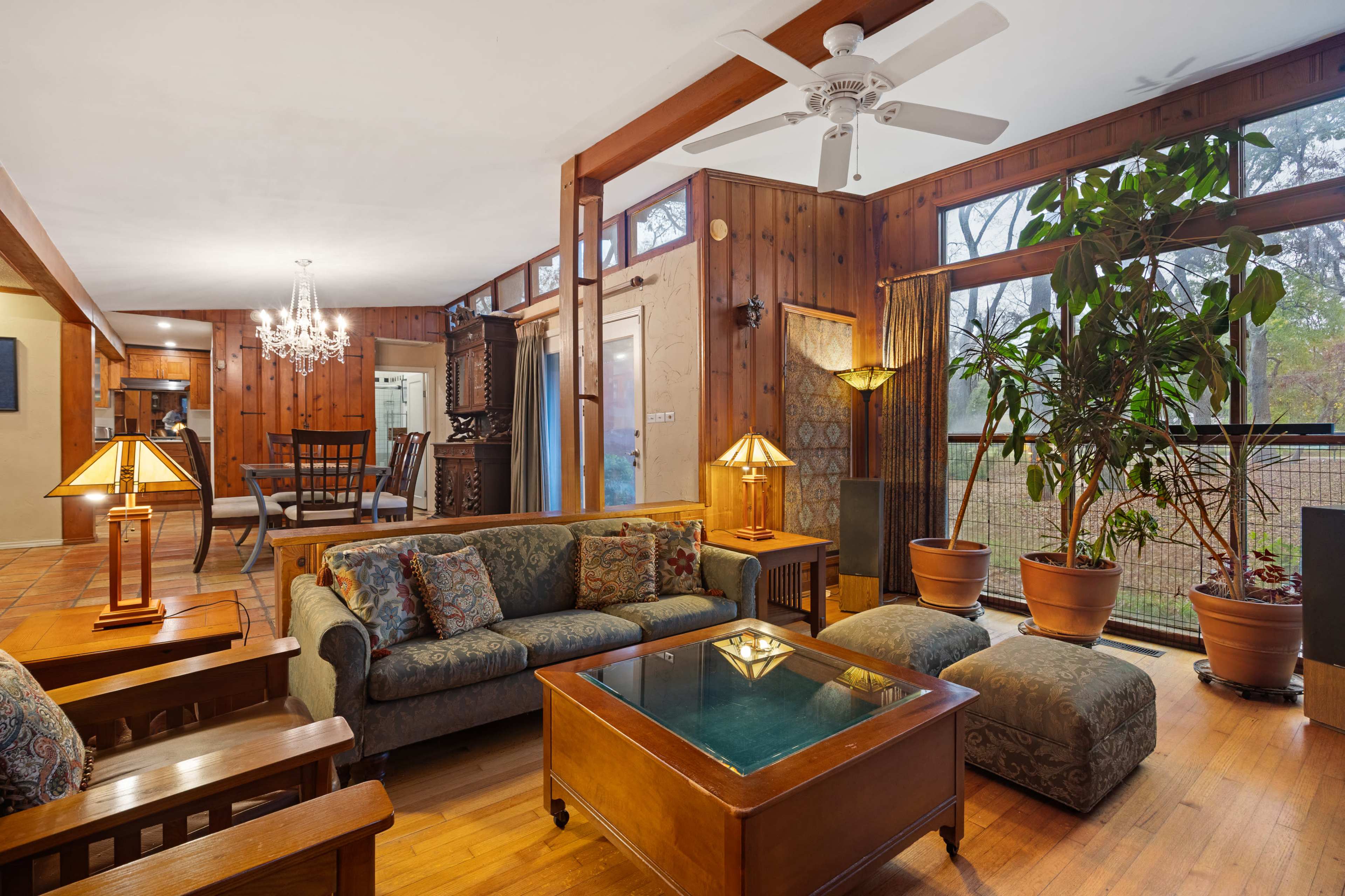 The image shows a cozy living room with wooden paneling, a comfortable seating area, and large windows filled with greenery.