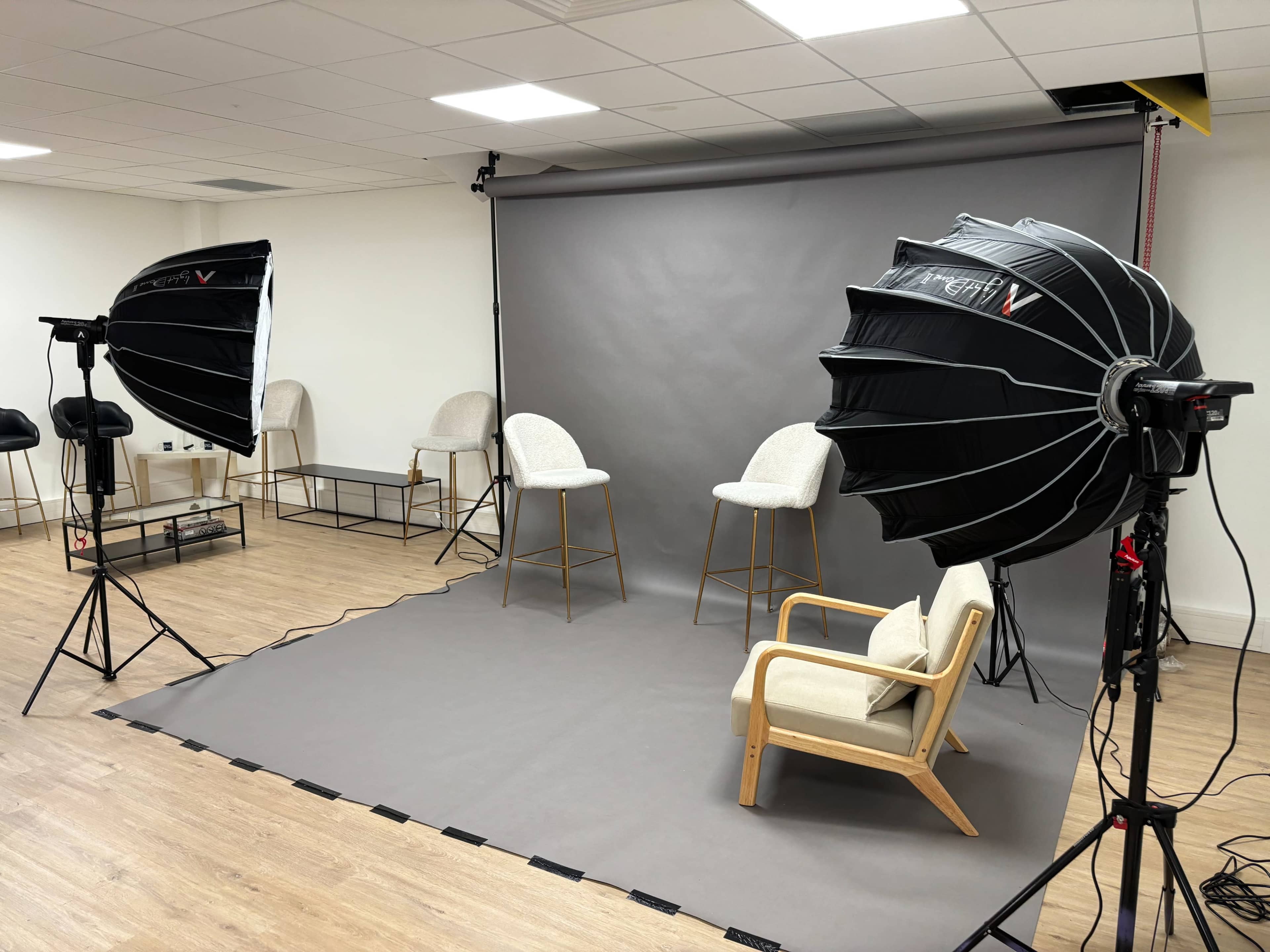 A photography studio setup featuring two softbox lights, a grey backdrop, and several high stools arranged around a wooden chair.