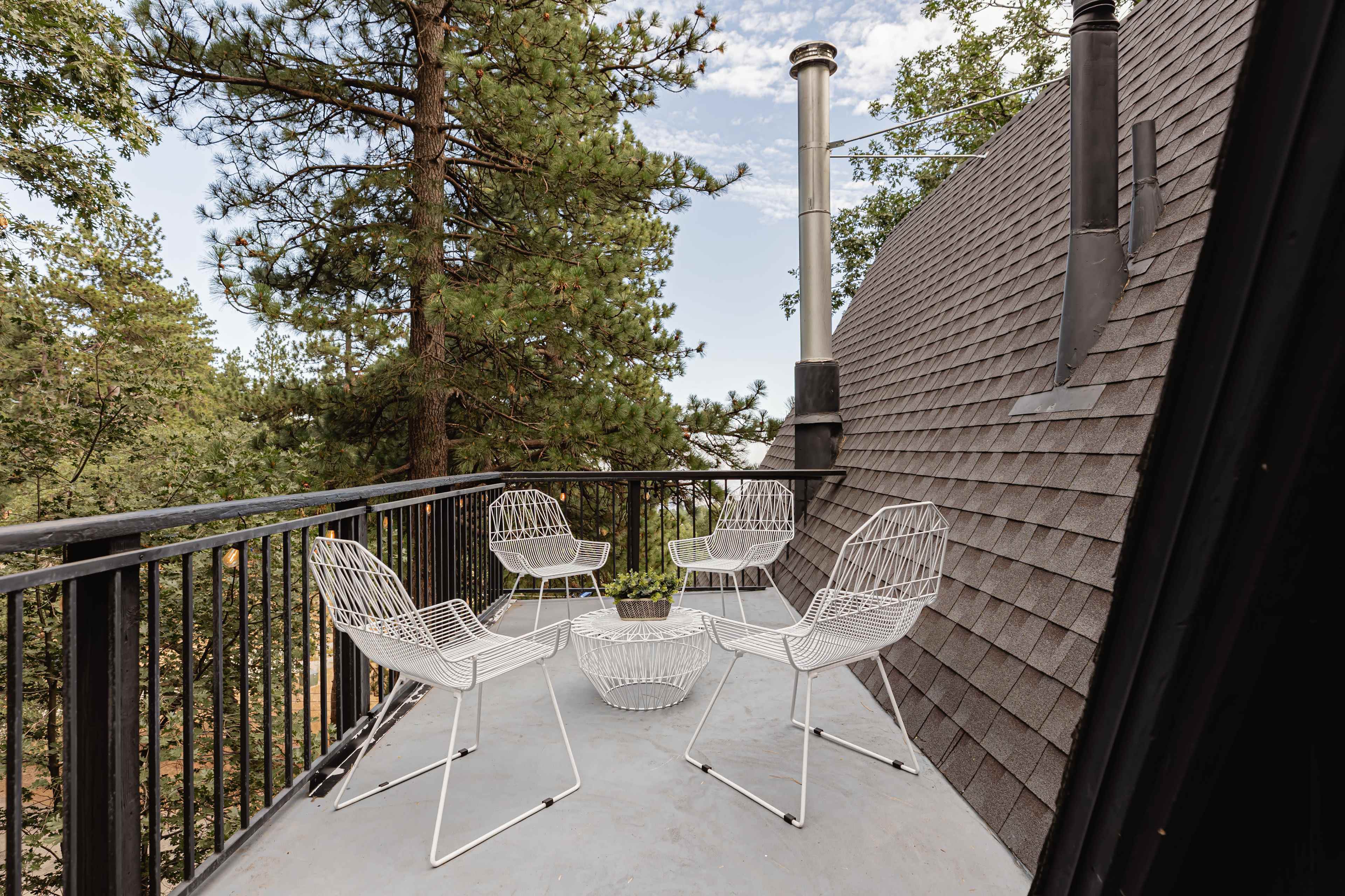 A balcony features five white wire chairs arranged around a circular wire table, surrounded by tall trees and a clear sky.