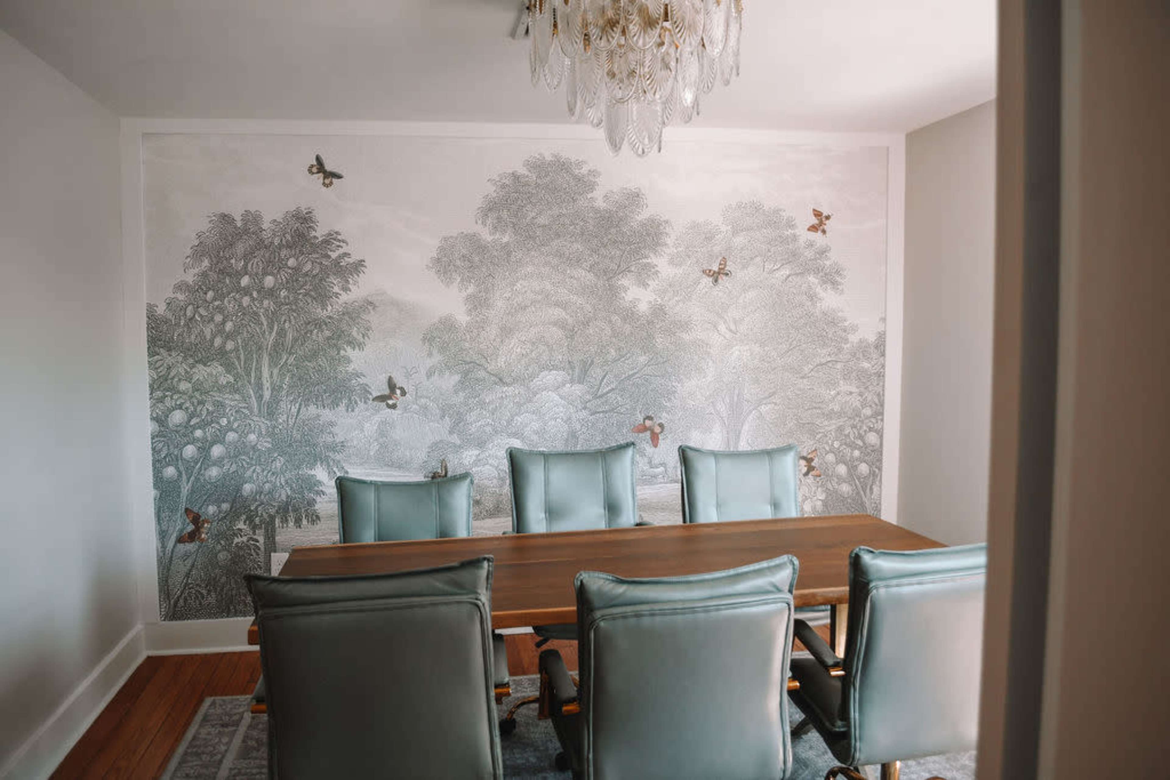 The image depicts a dining room with a large wooden table surrounded by six teal chairs and a detailed mural of trees and birds on the wall.