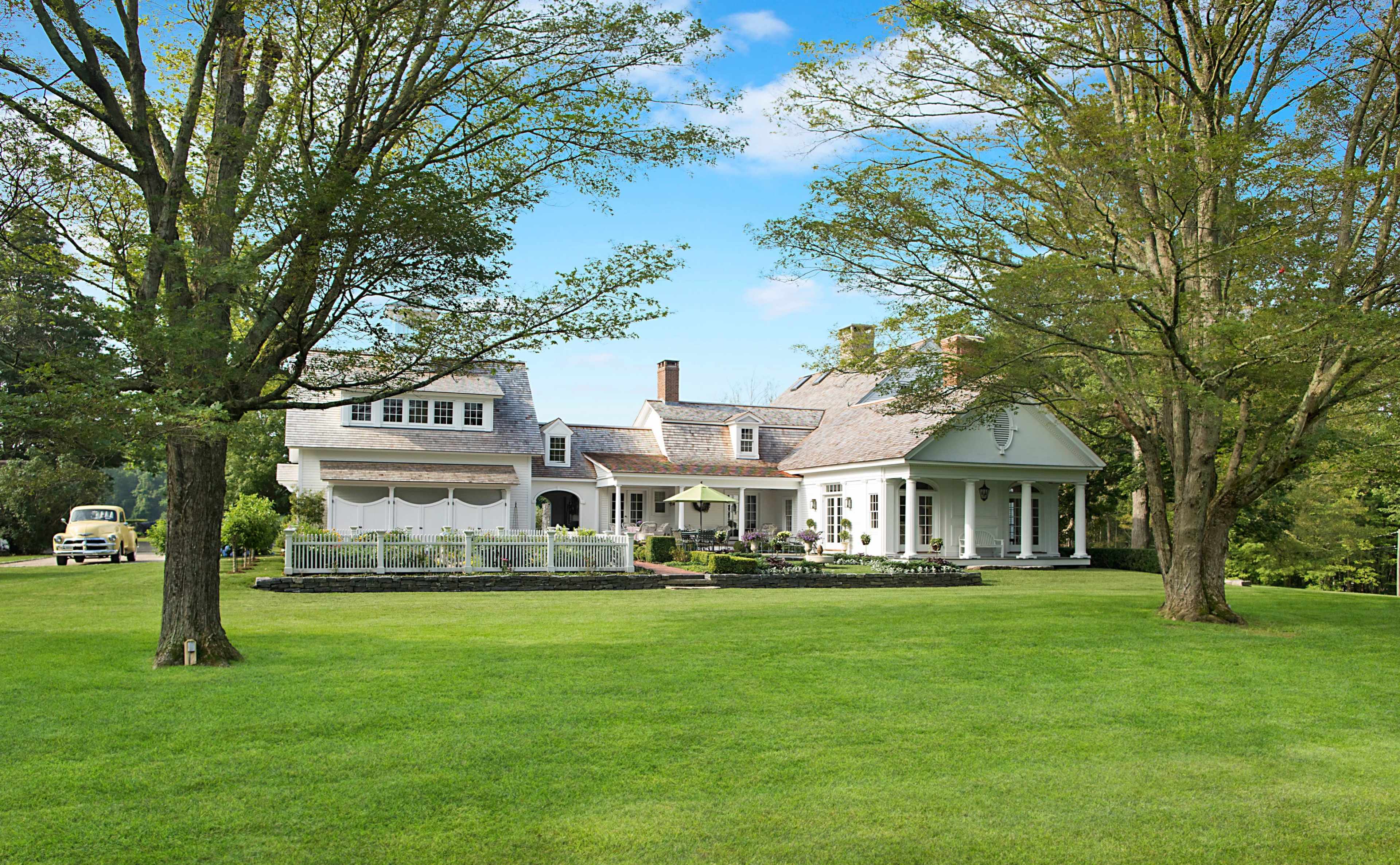 A large, white two-story house is surrounded by a lush green lawn and tall trees, with a stone walkway leading to the entrance.