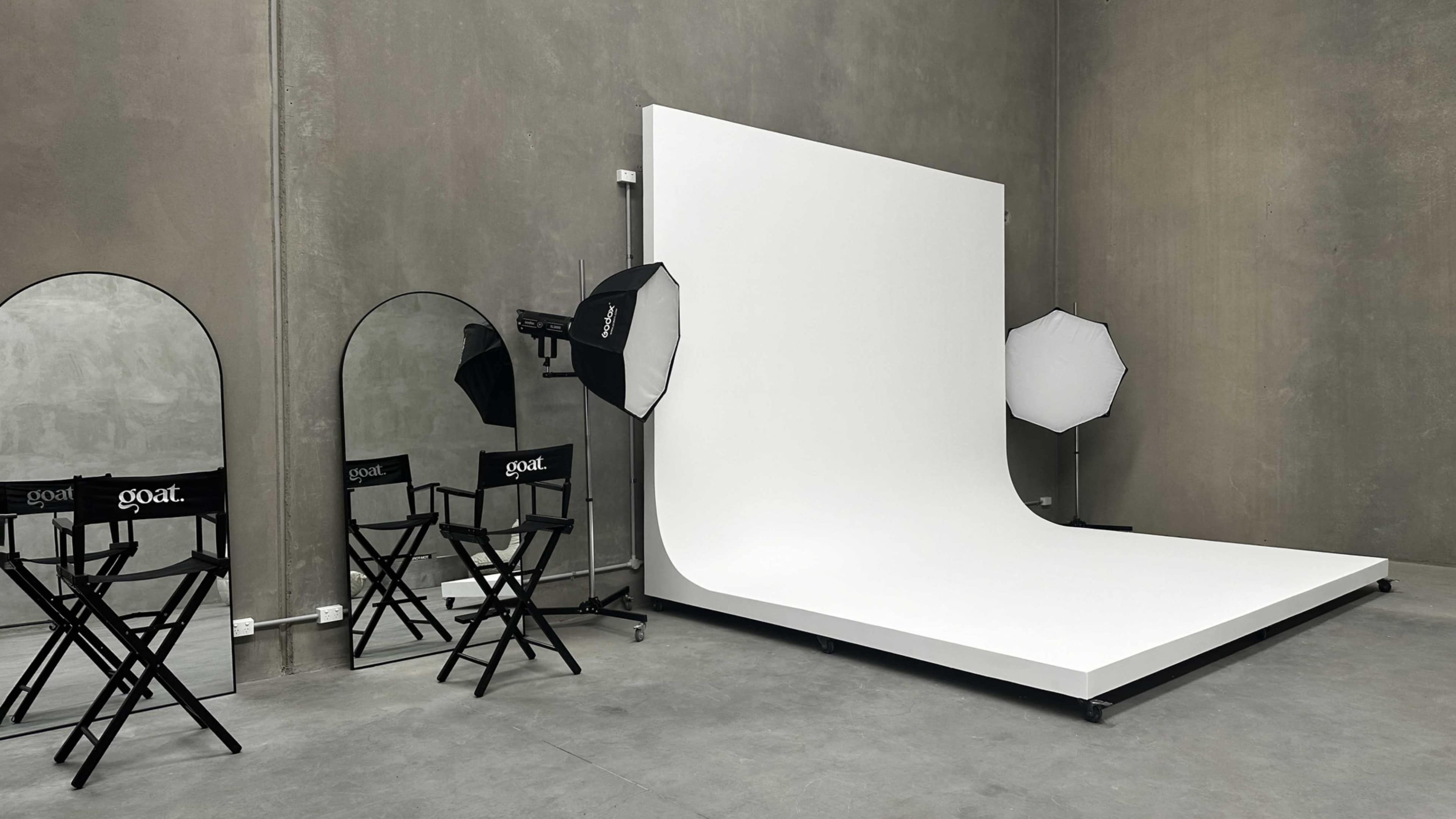 A minimalist photography studio with a white backdrop, two softbox lights, and two director's chairs labeled "goat."
