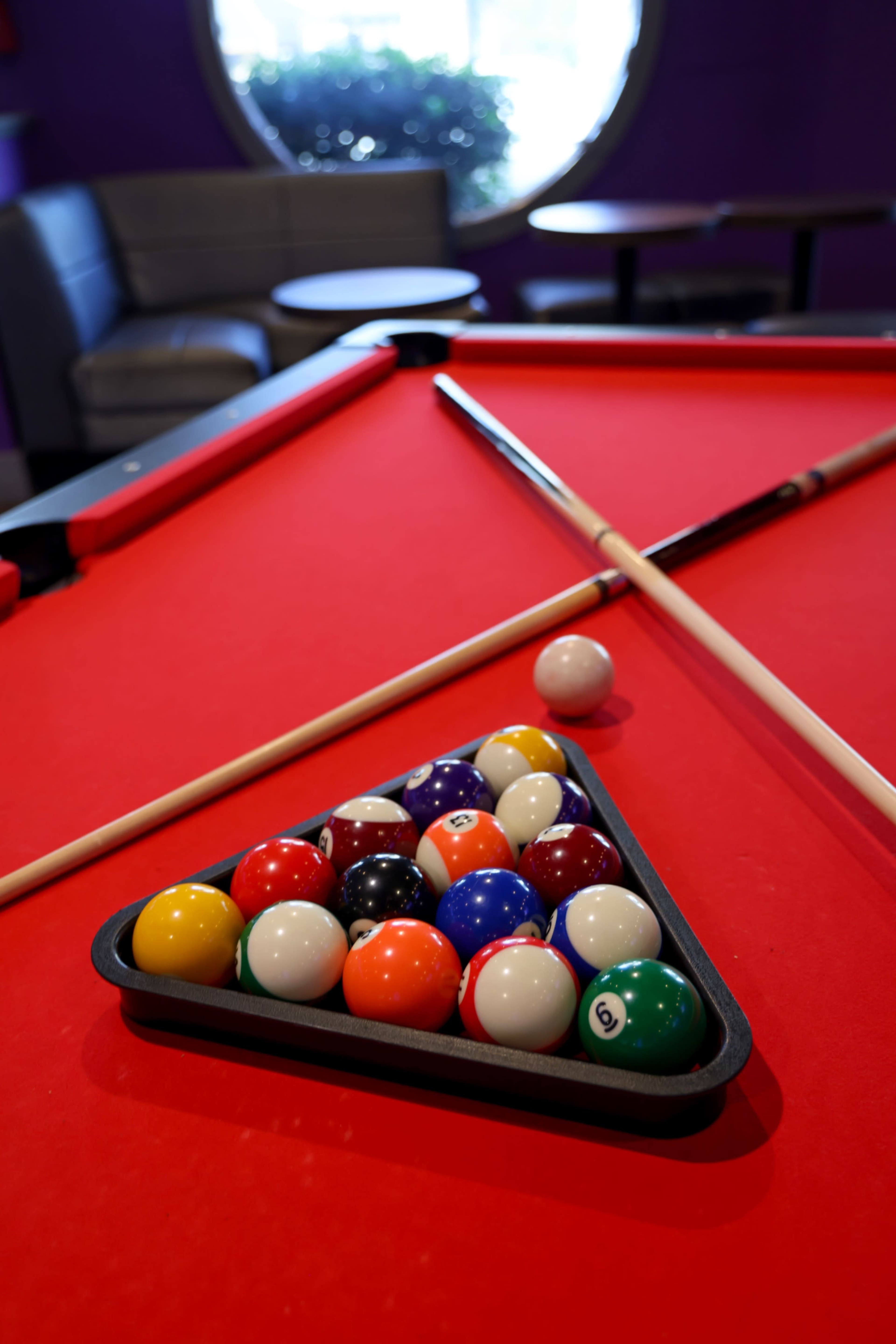 A triangular rack of billiard balls sits on a red pool table, with two cues crossed over it and a single cue ball nearby.