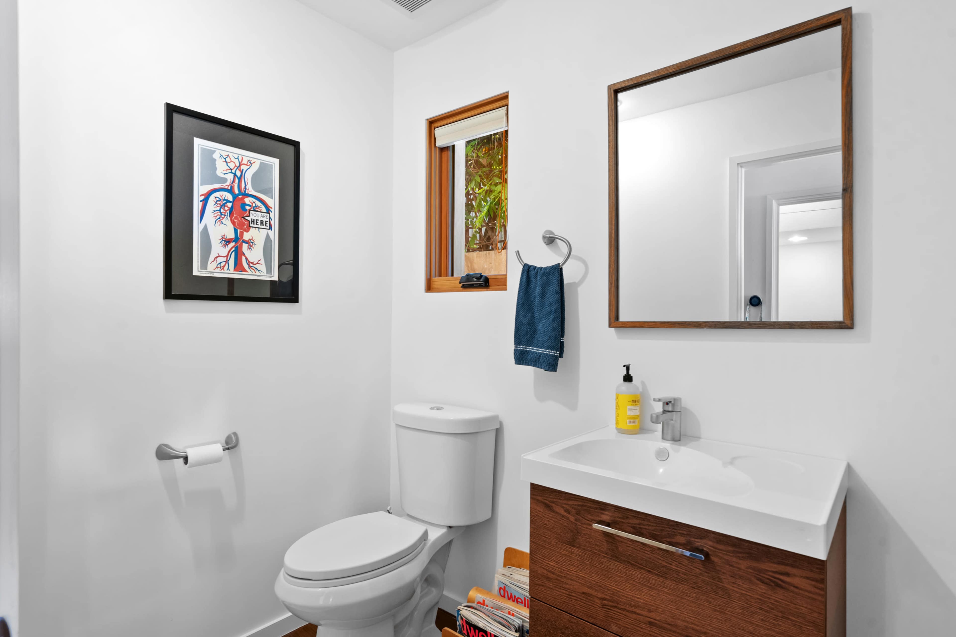 A modern bathroom featuring a toilet, sink with a wooden cabinet, a mirror, and a framed anatomical heart illustration on the wall.