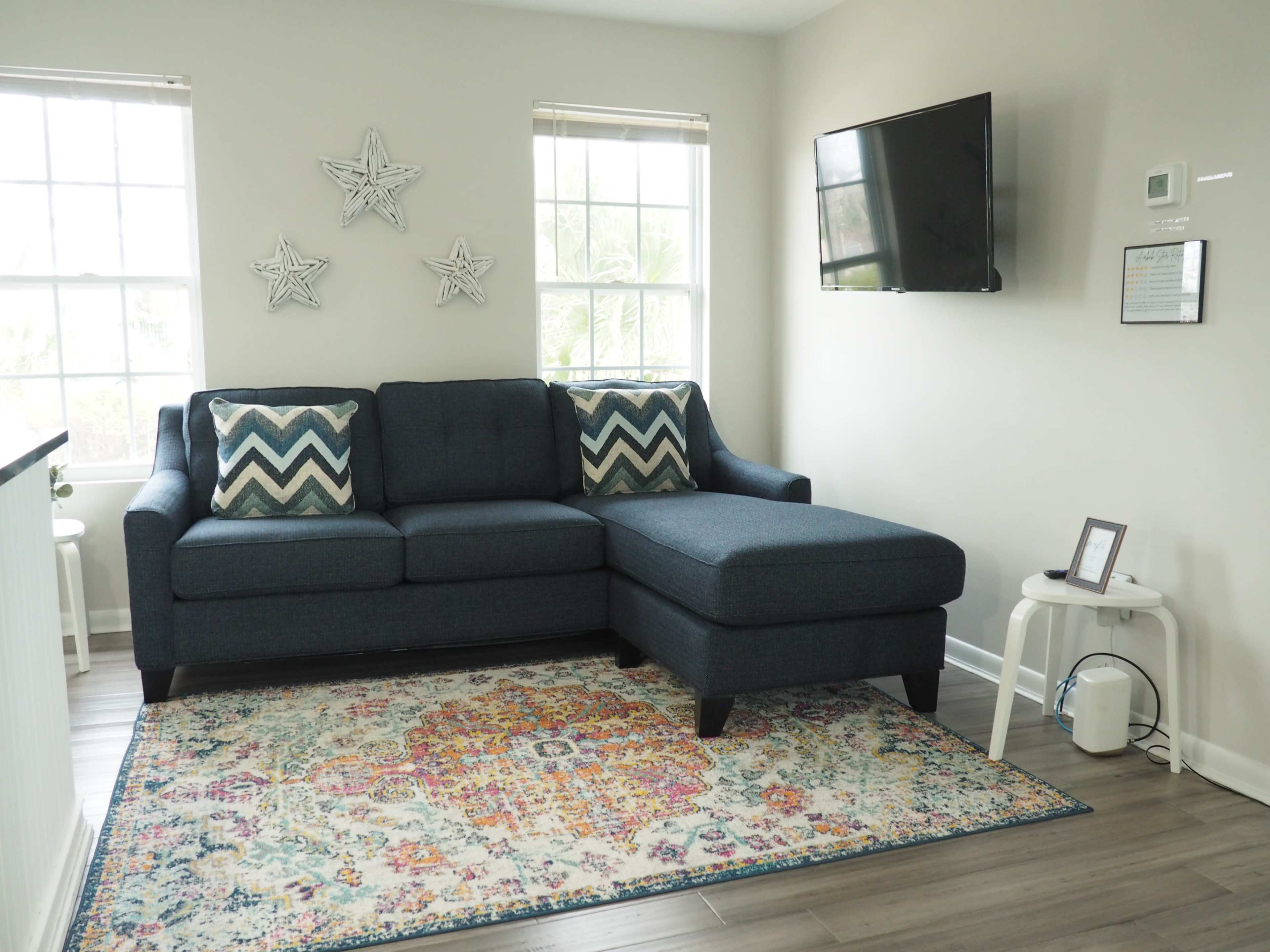 A living room with a blue sectional sofa, decorative star wall hangings, a television mounted on the wall, and a colorful area rug on the floor.
