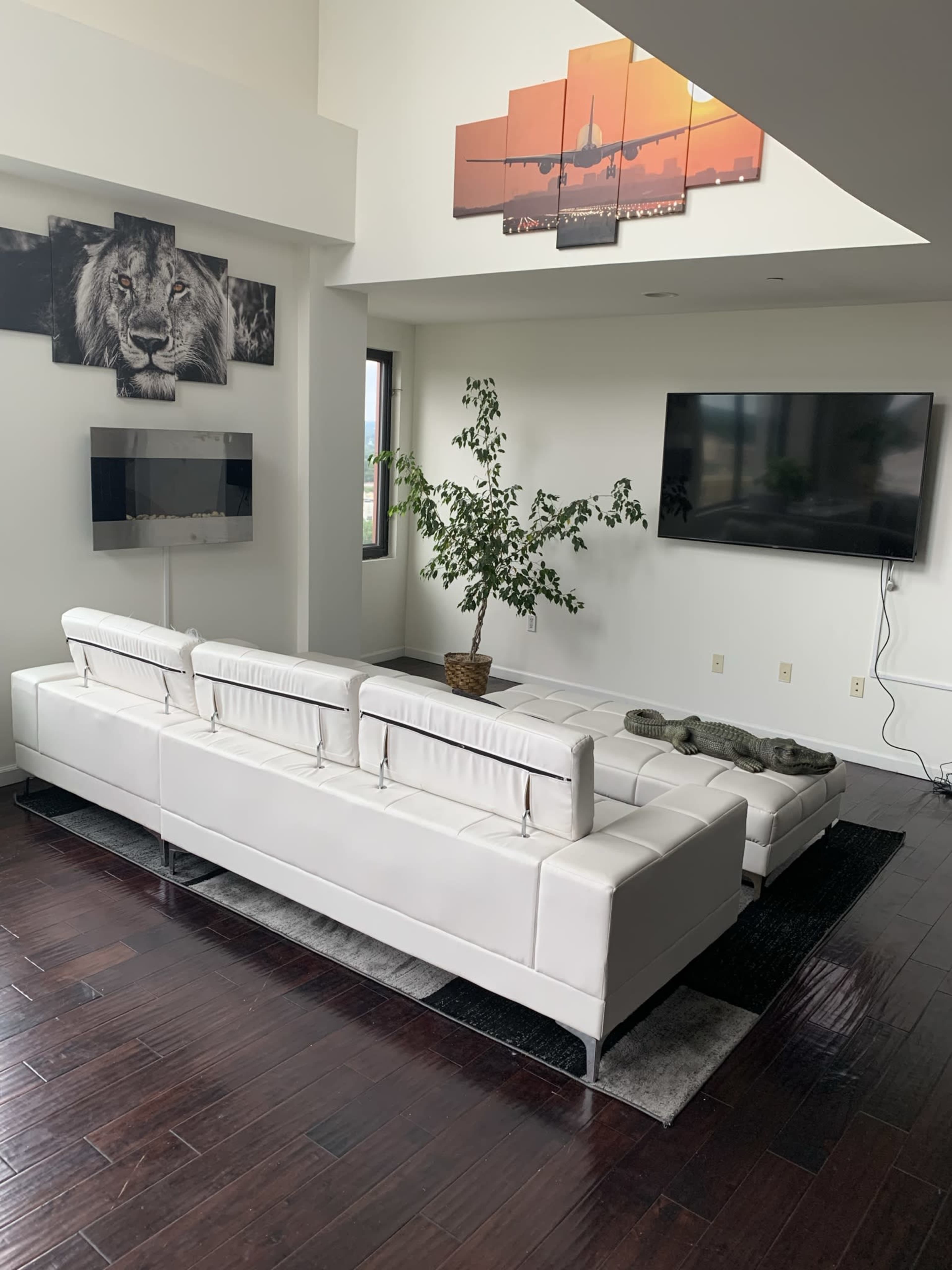 A modern living room features a white sectional sofa, a potted plant, and two wall-mounted televisions with a large framed photograph above the sofa.