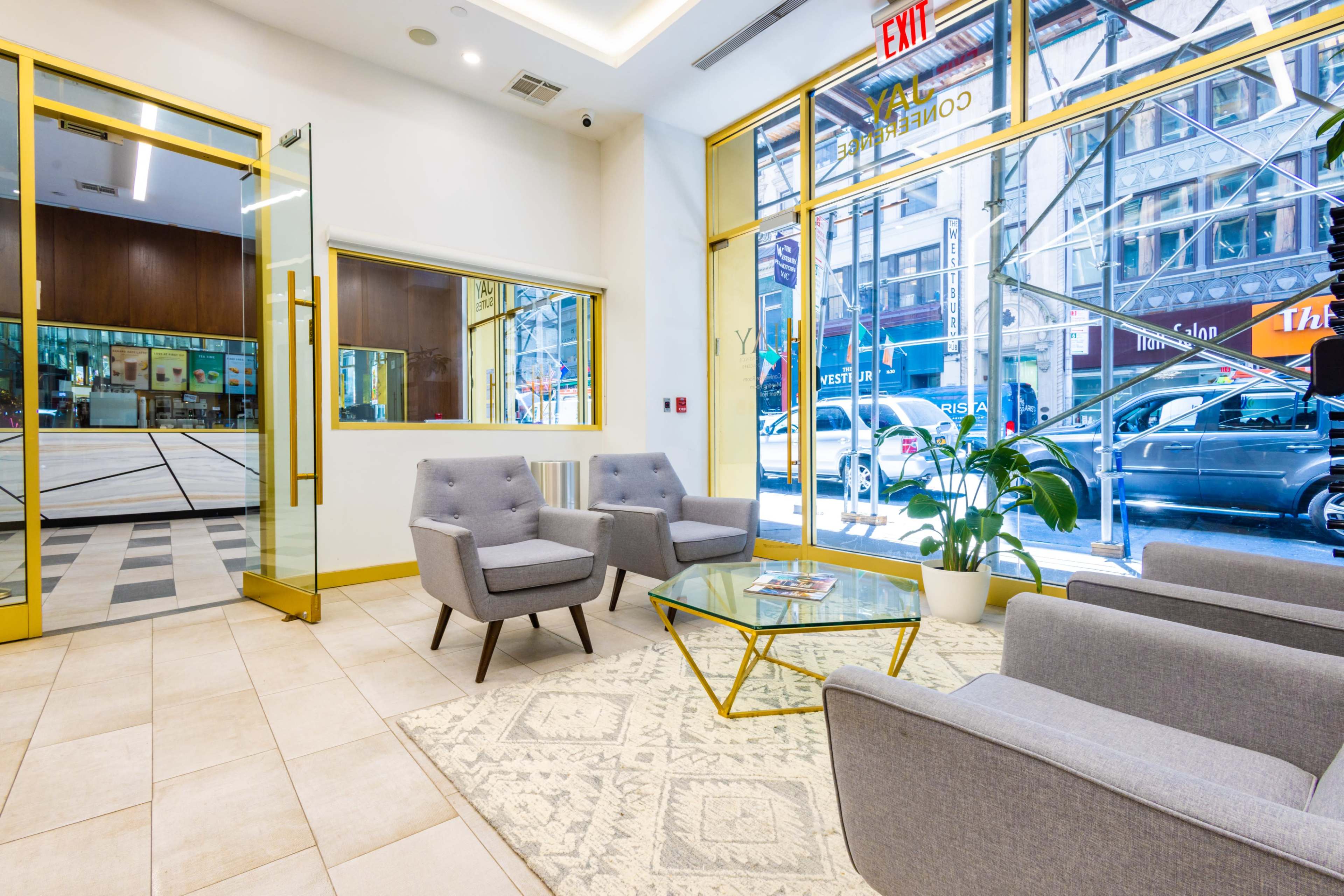 The image shows a modern lobby with gray armchairs, a glass coffee table, and large windows overlooking a city street.
