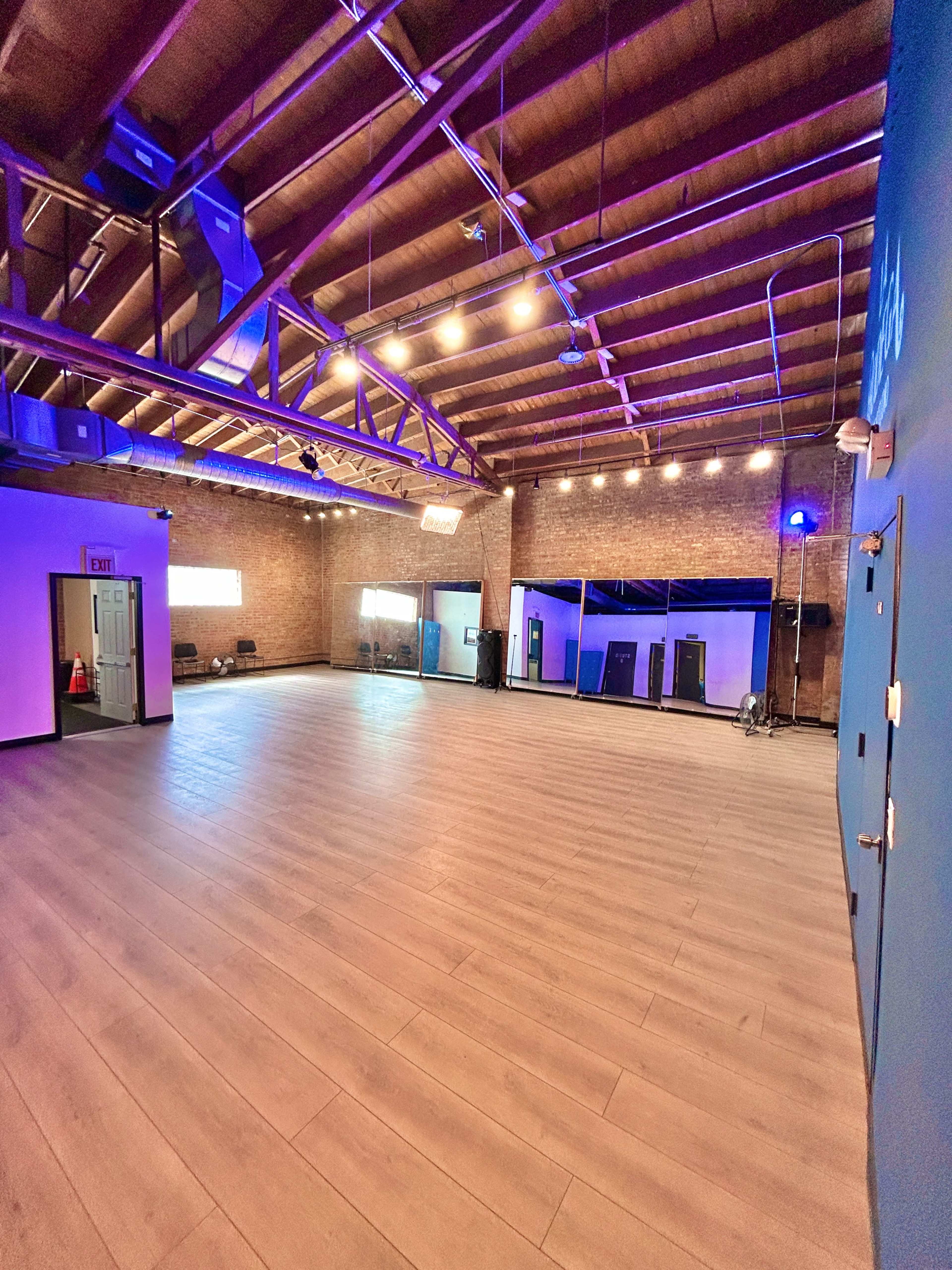 The image shows a spacious, well-lit room with high wooden ceilings, wooden flooring, and brick walls, equipped with a few chairs and large mirrors.