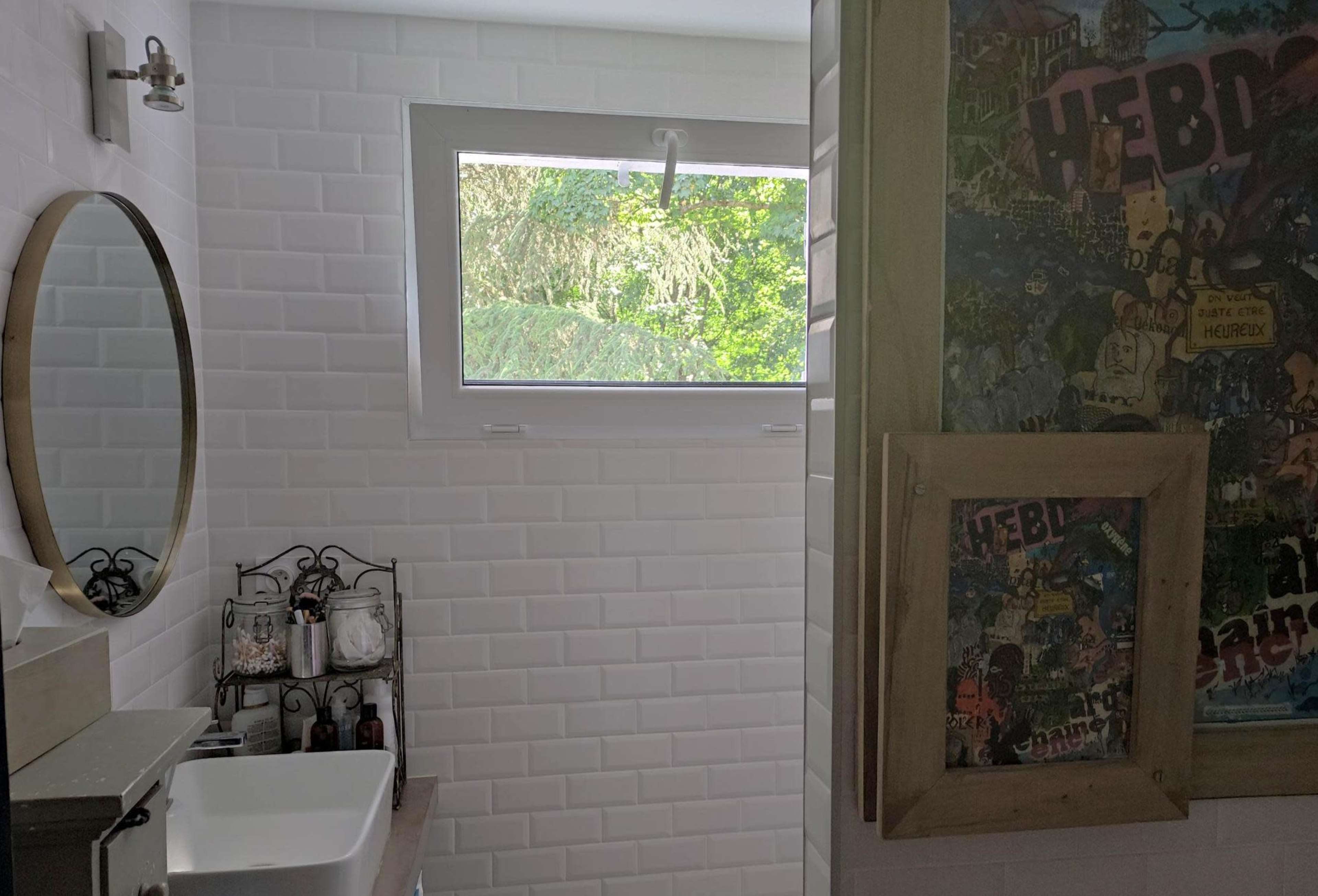 A bathroom with white tiled walls, a circular mirror, a sink, and a window providing natural light.