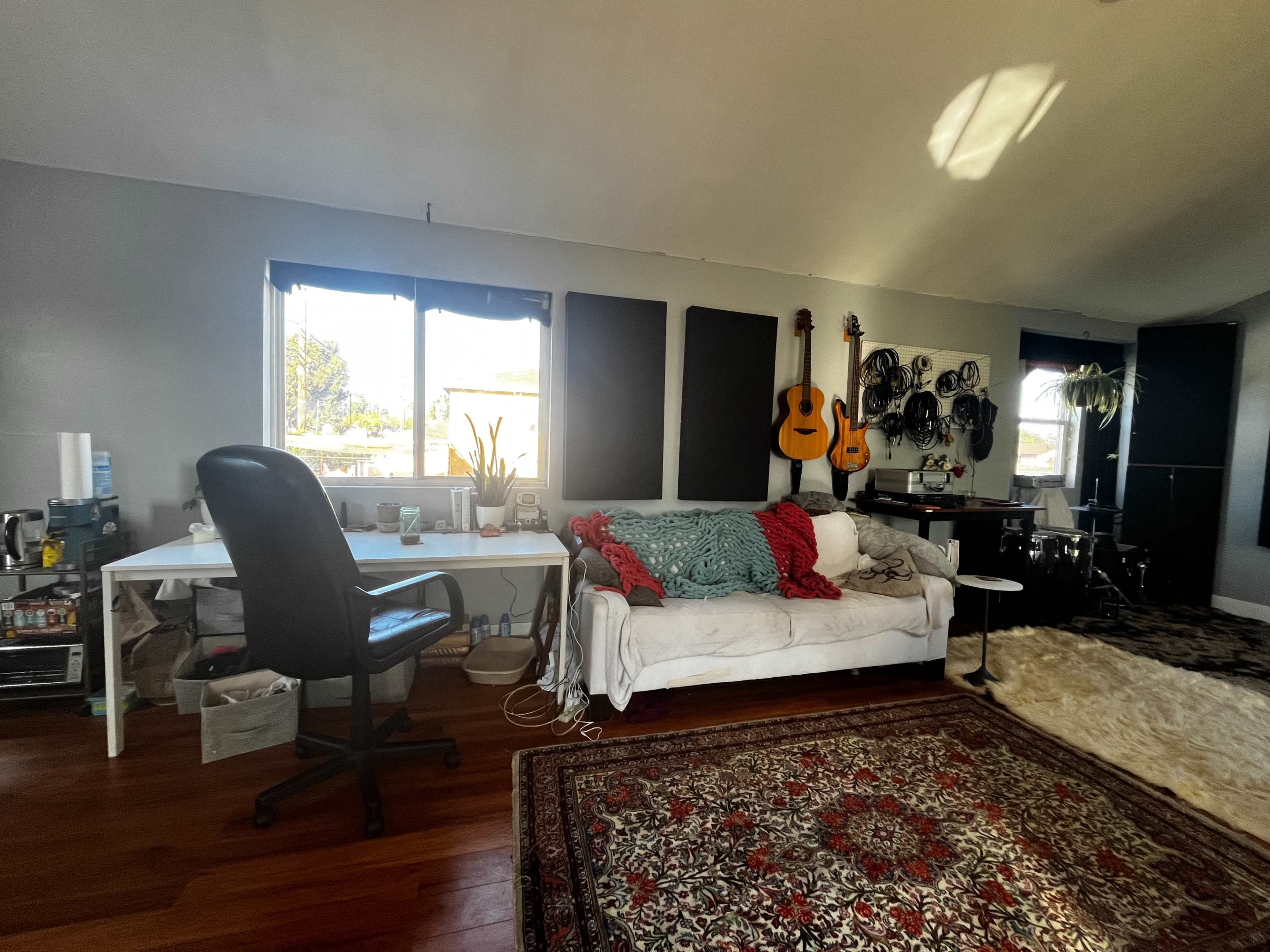 The image shows a spacious room with a white couch, a workspace with a desk and a chair, and musical instruments displayed on the wall.
