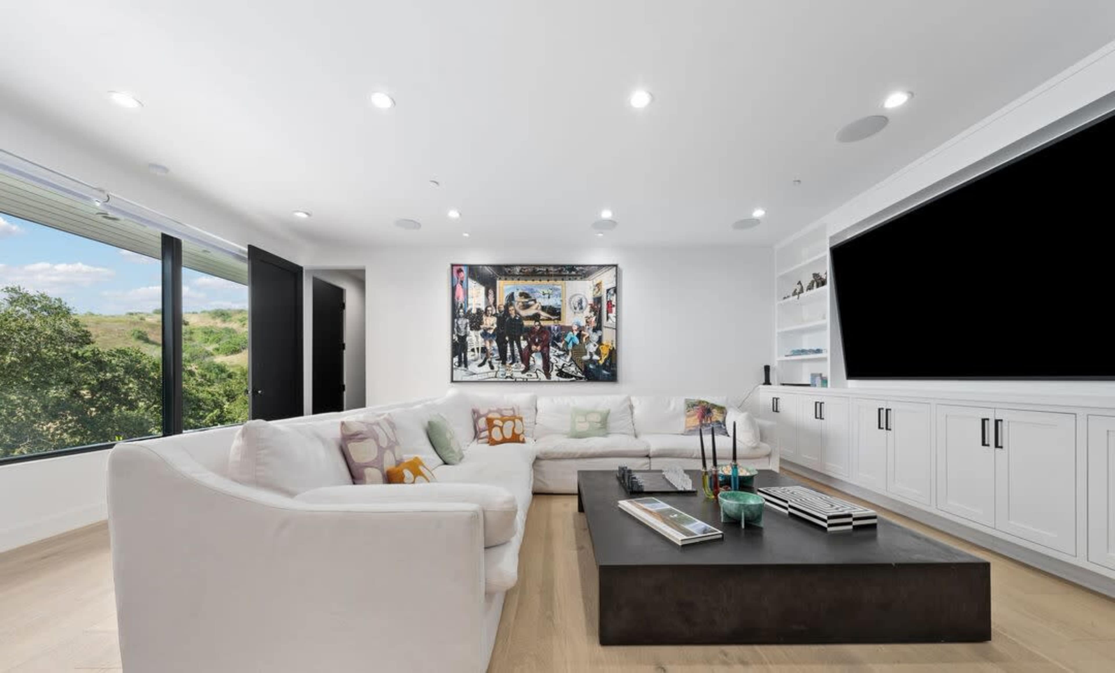 The room features a large white sectional sofa, a dark coffee table, a mounted flat-screen TV, and a prominent wall artwork, along with large windows overlooking greenery.