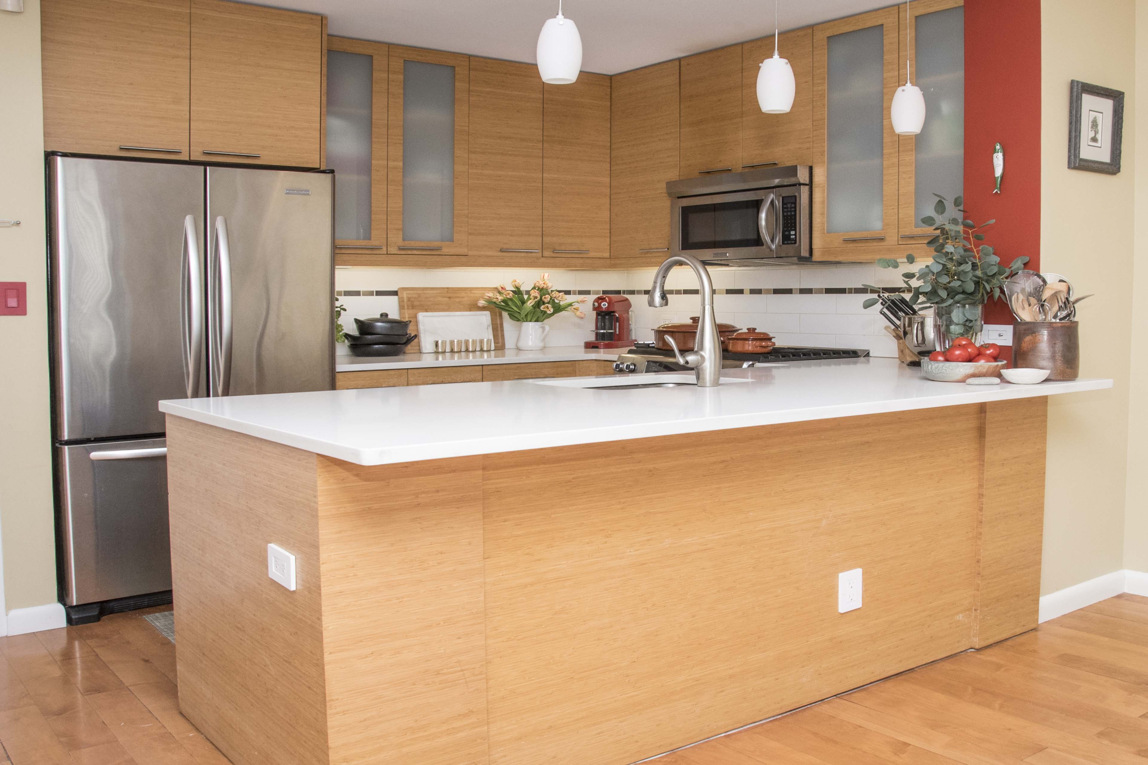 The image shows a modern kitchen featuring bamboo cabinets, stainless steel appliances, and a large island with a white countertop.