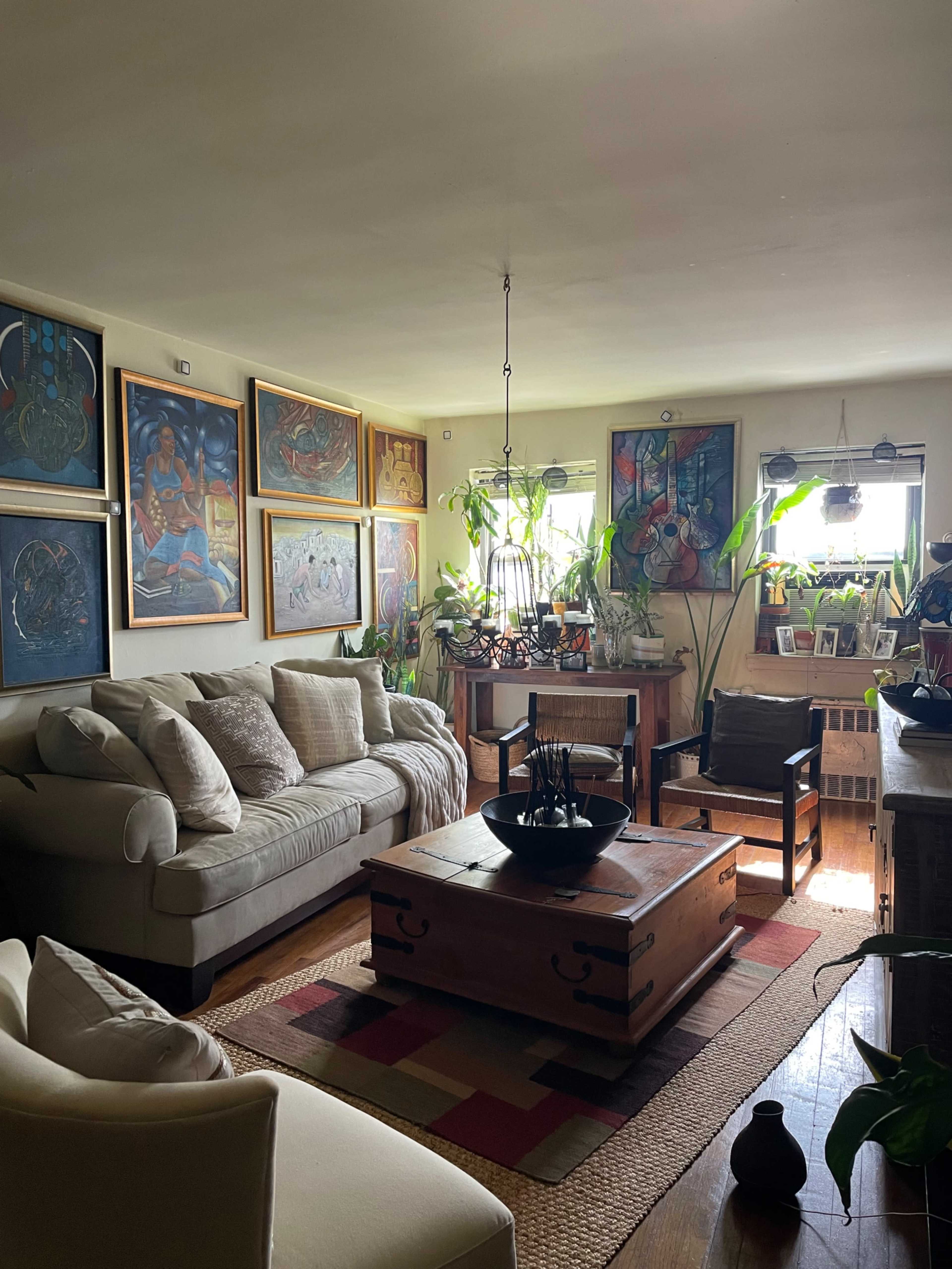 A living room features a beige sofa, a wooden coffee table, and several framed artworks on the walls, with various plants placed around the space.