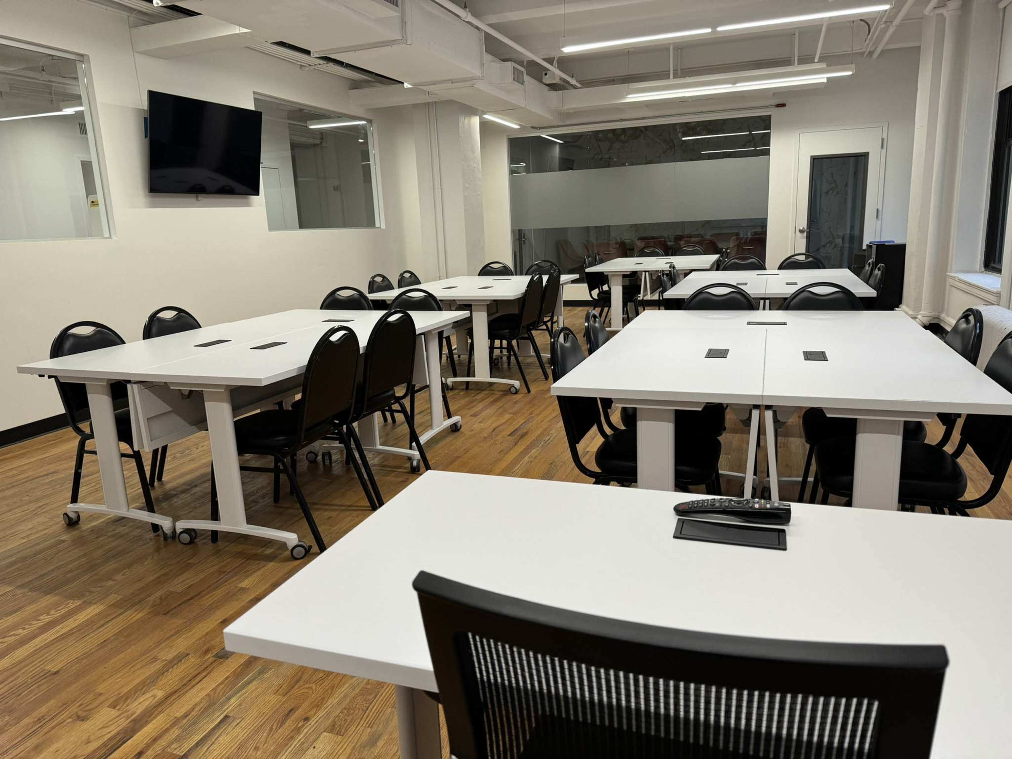 The image shows a modern classroom or meeting space with several white tables and black chairs arranged neatly on wooden flooring.