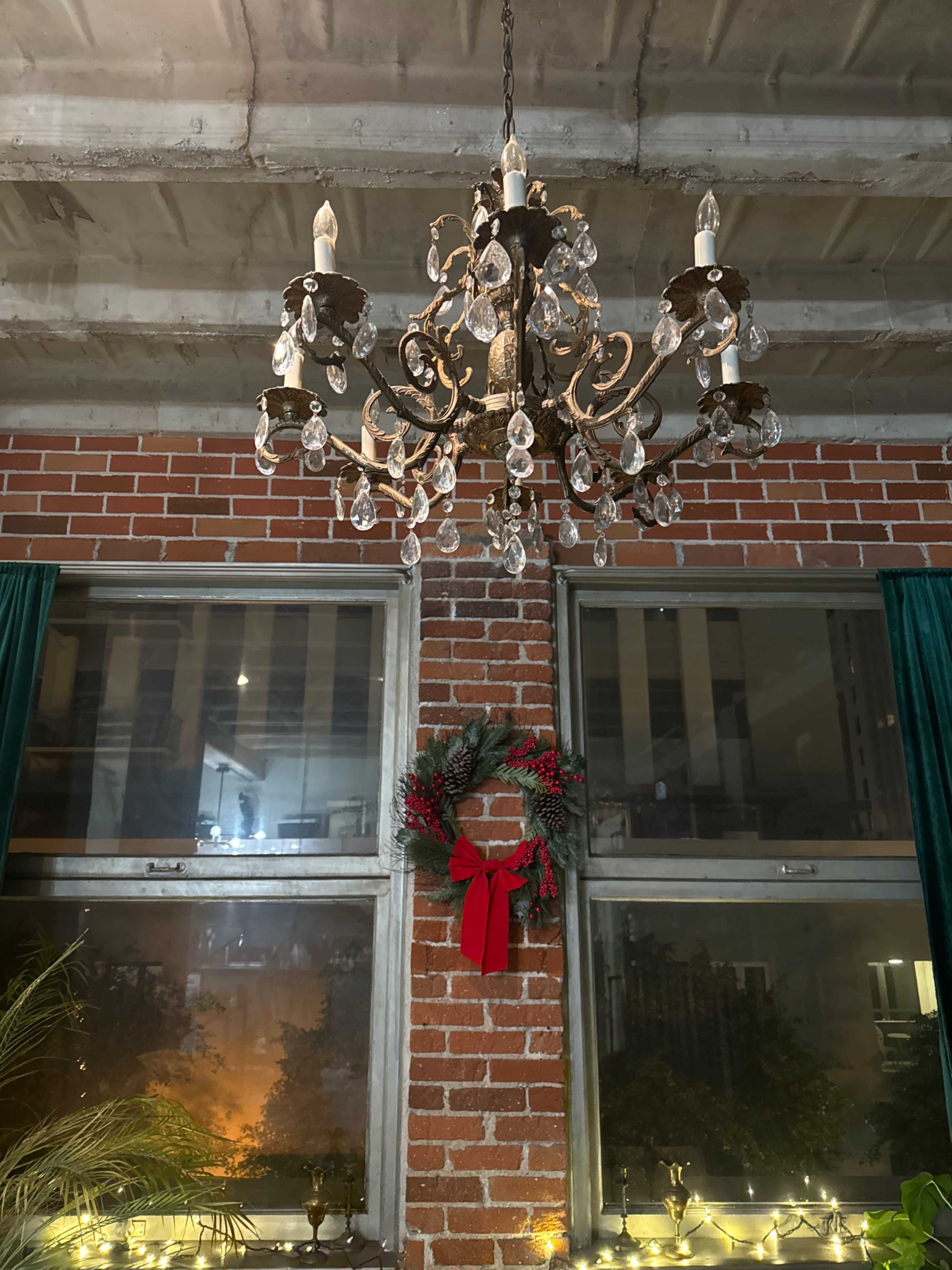 A decorative chandelier hangs from the ceiling above a red brick wall, with a holiday wreath adorned with a bow positioned between two windows.