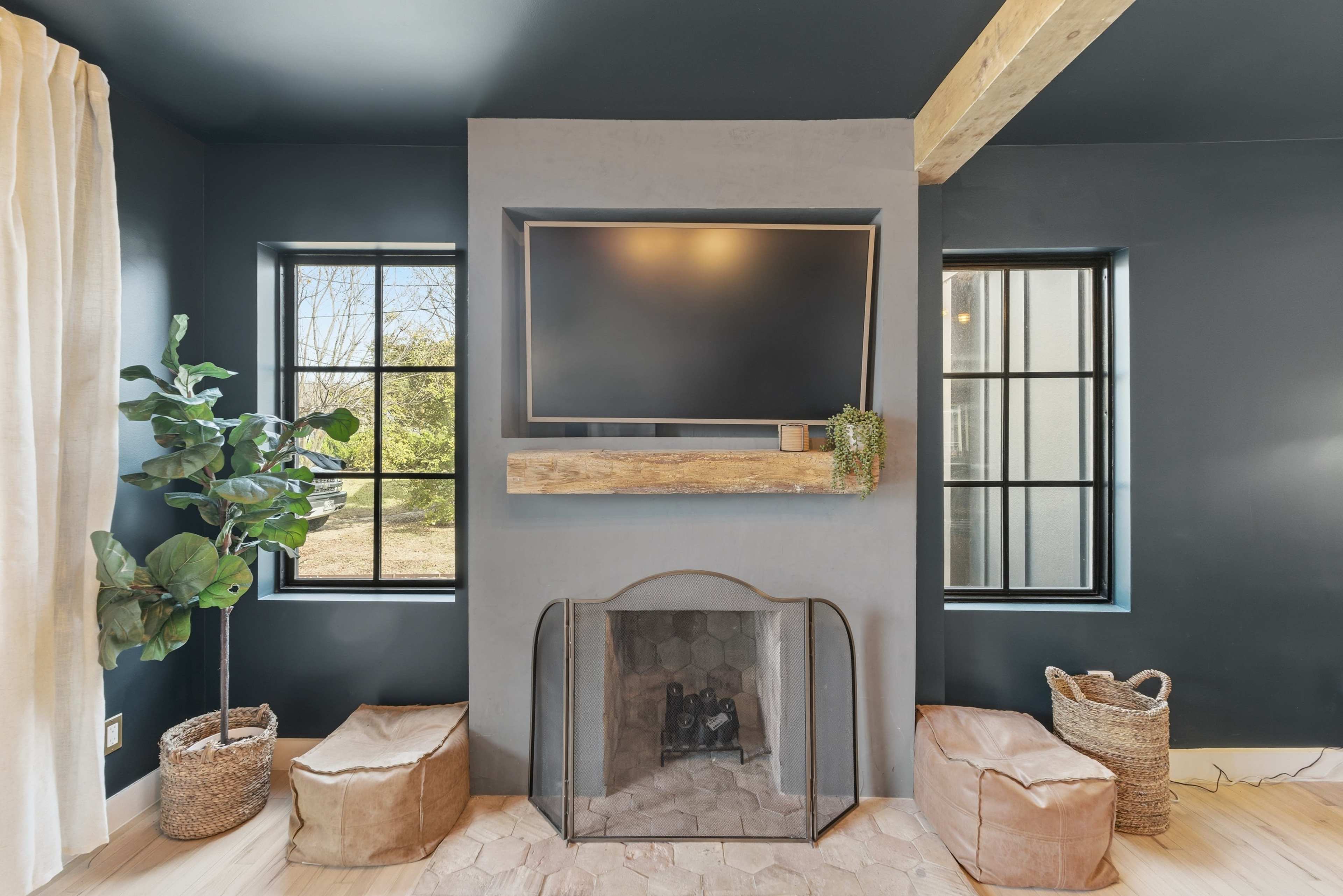 The image features a modern living room with a mounted television above a wooden shelf and a fireplace, flanked by two windows and styled with indoor plants and brown poufs.